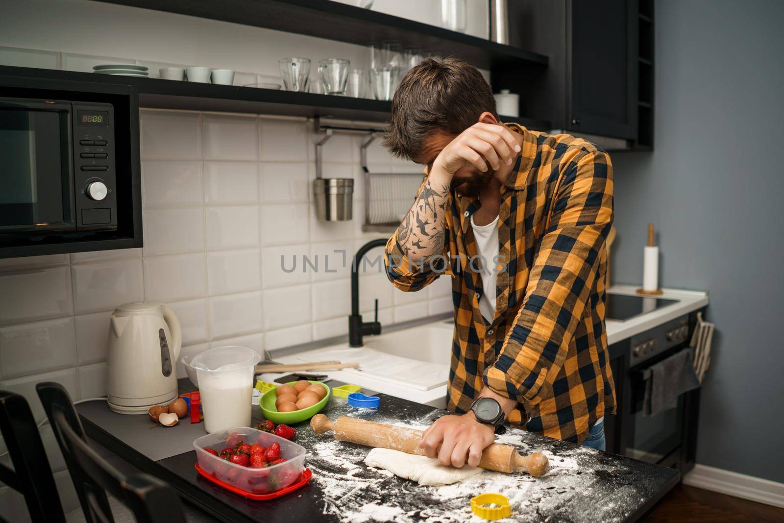 Young man is making cookies in his kitchen. He is tired and bored.