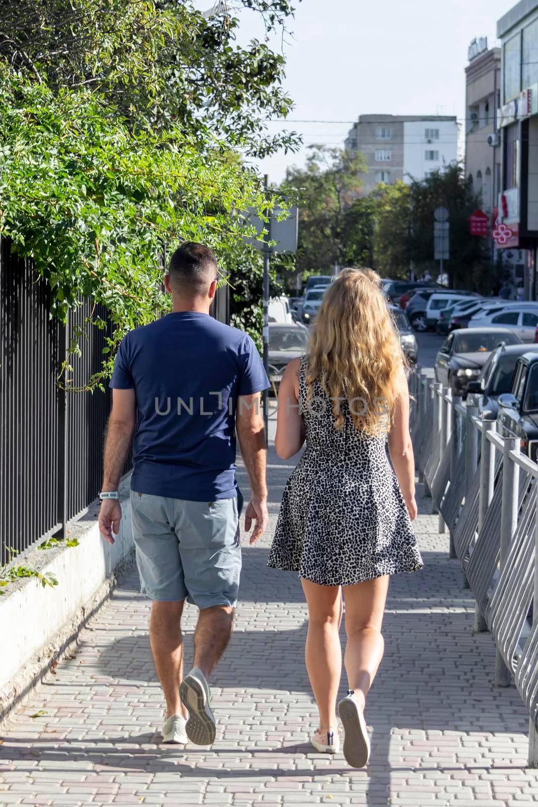 Man and woman pair walk along pavement along road. Sunny summer day. Crimea, Sudak - 10 October 2020. by Essffes