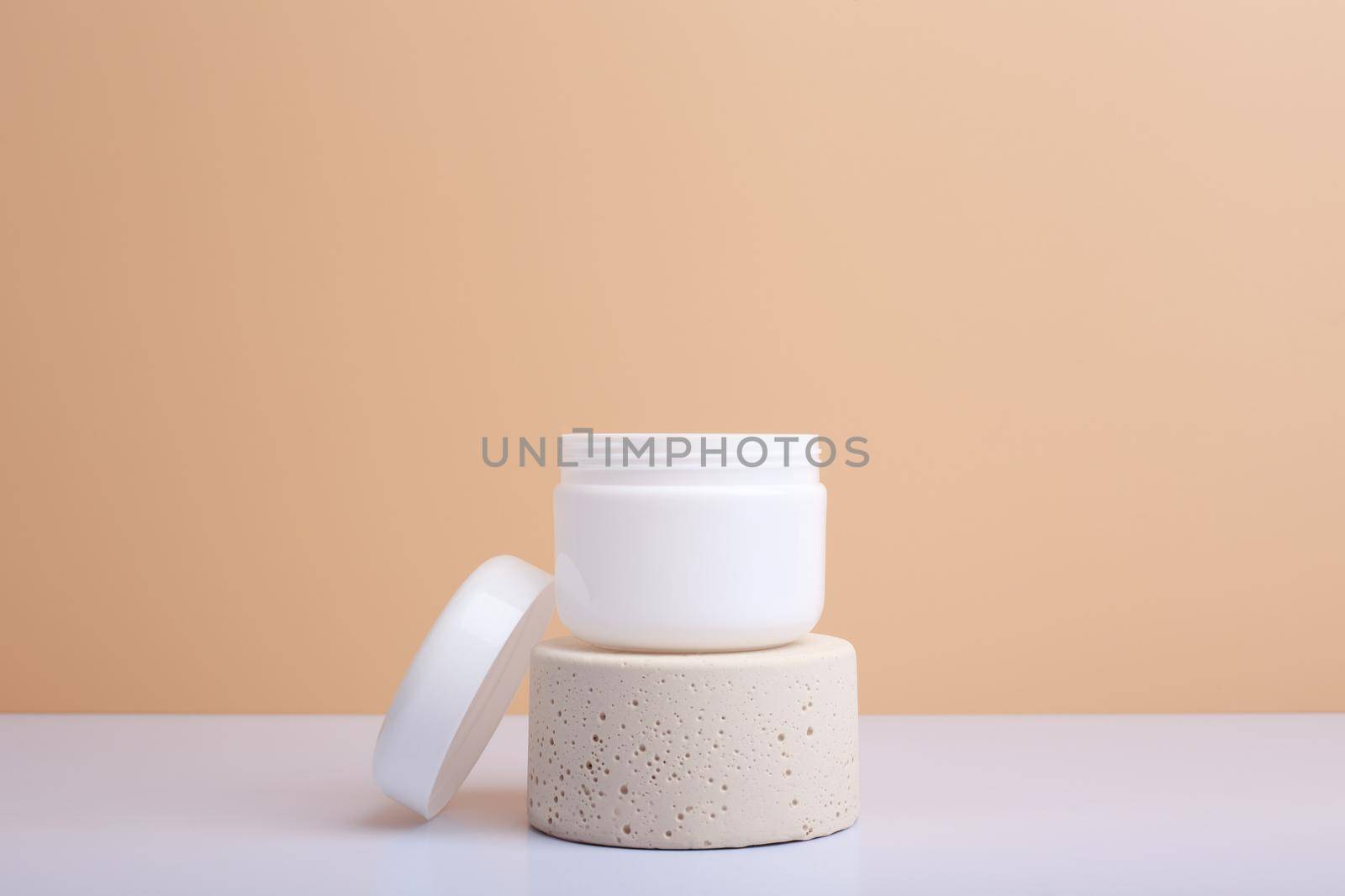 White opened cosmetic jar on beige podium and white table against beige background with copy space. Concept of beauty products for skin. Opened jar with balm, lotion, cream, scrub or mask.