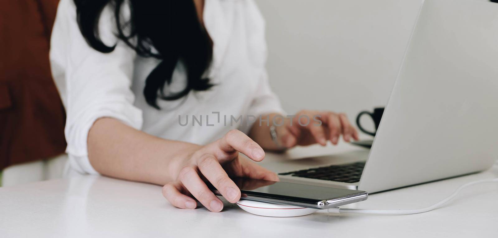 Cropped image of an office woman is charging a smartphone with a wireless charger while typing on a computer laptop. by wichayada