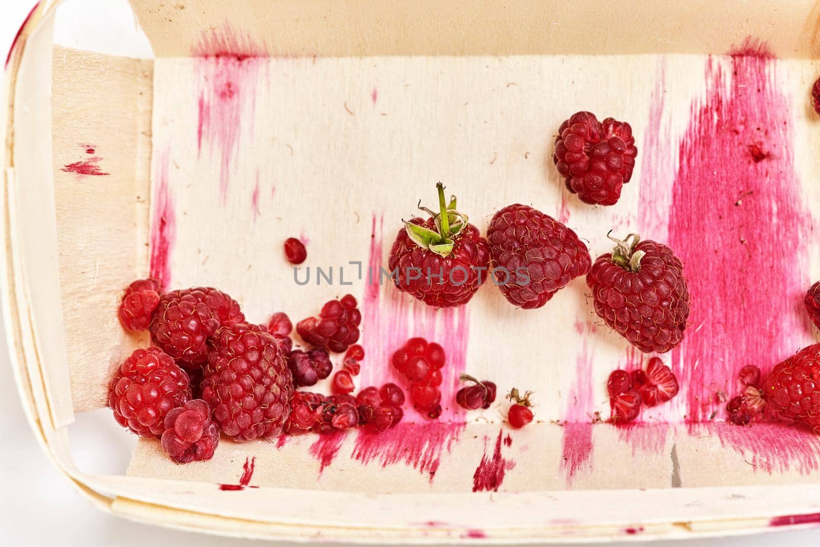 The half-eaten remnants of ripe raspberries in a small wooden container with streaks of red juice. Top view close up