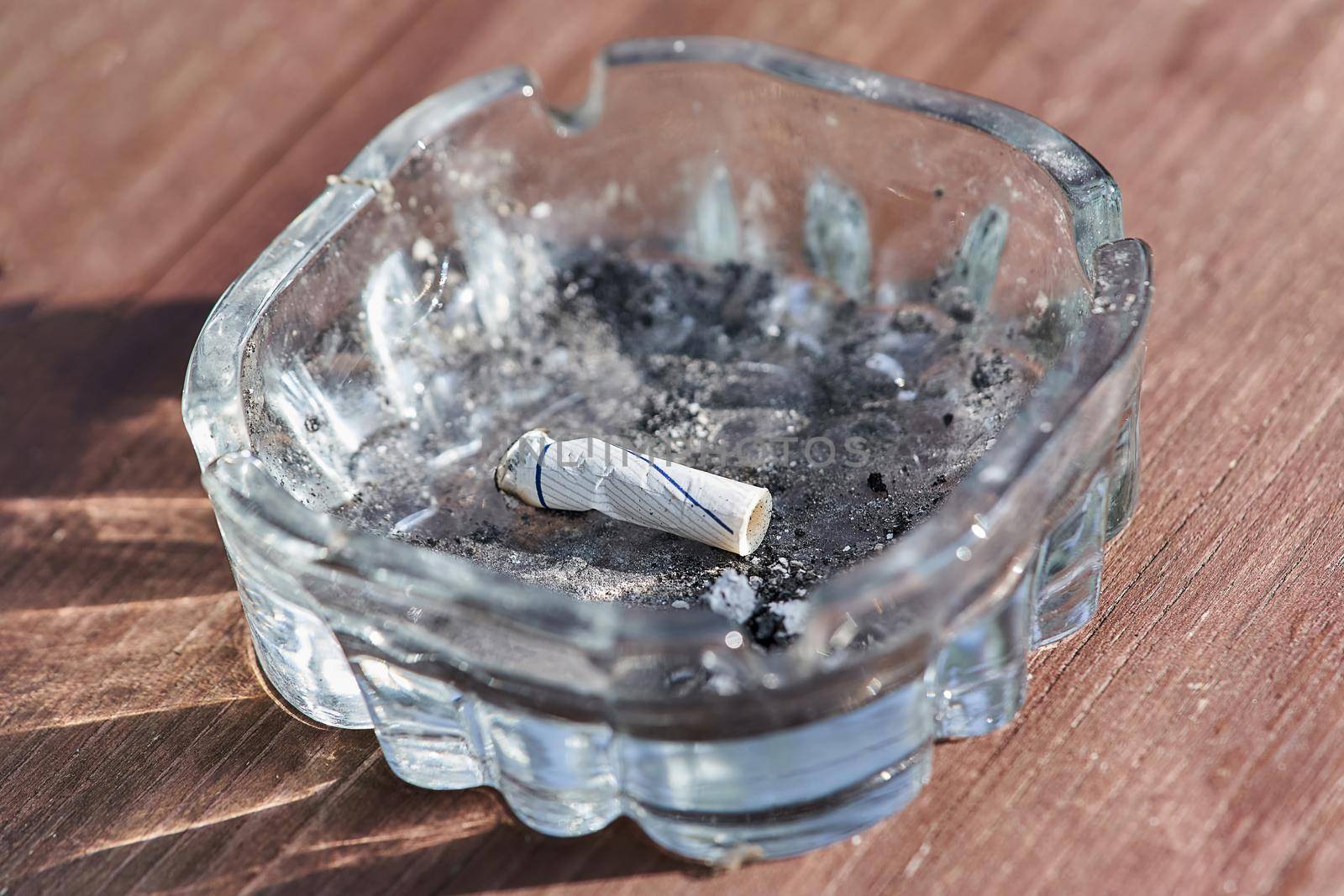 The cigarette butt lies in a dirty glass ashtray. Top view of brown planks background
