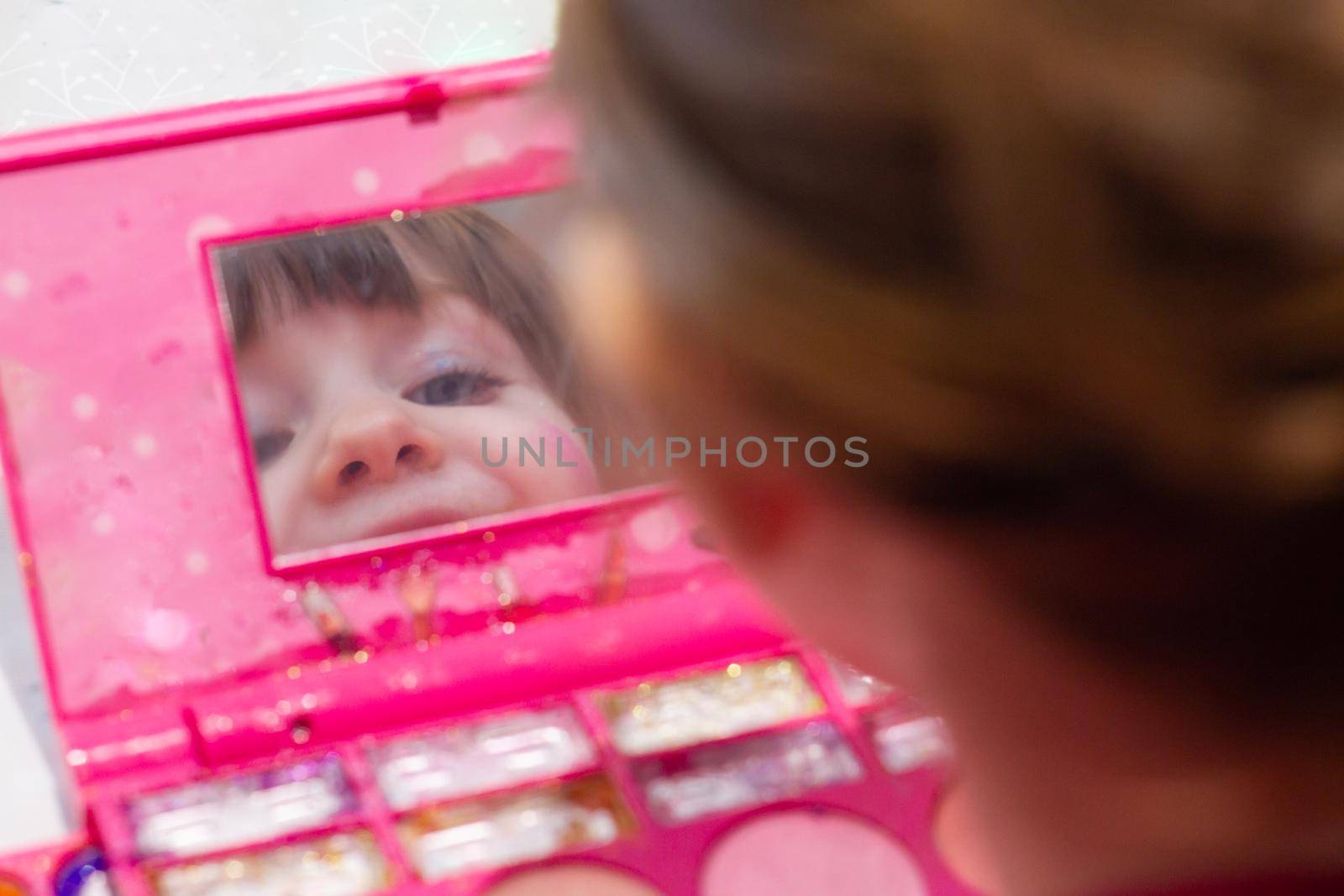 A cute blue-eyed girl with makeup in her face smiling and looking at a mirror in a makeup set