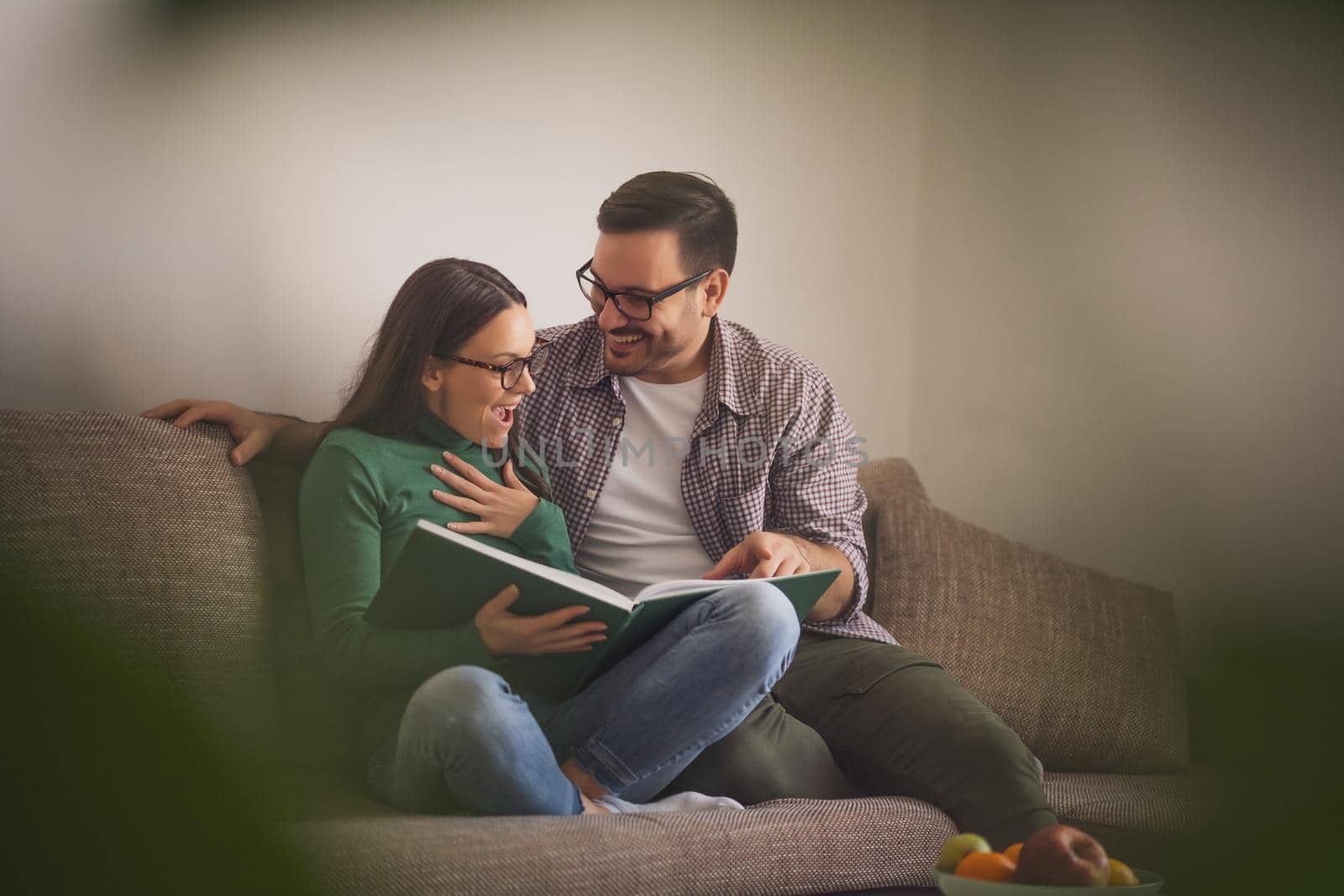 Happy couple is sitting at sofa in their home and looking at book.