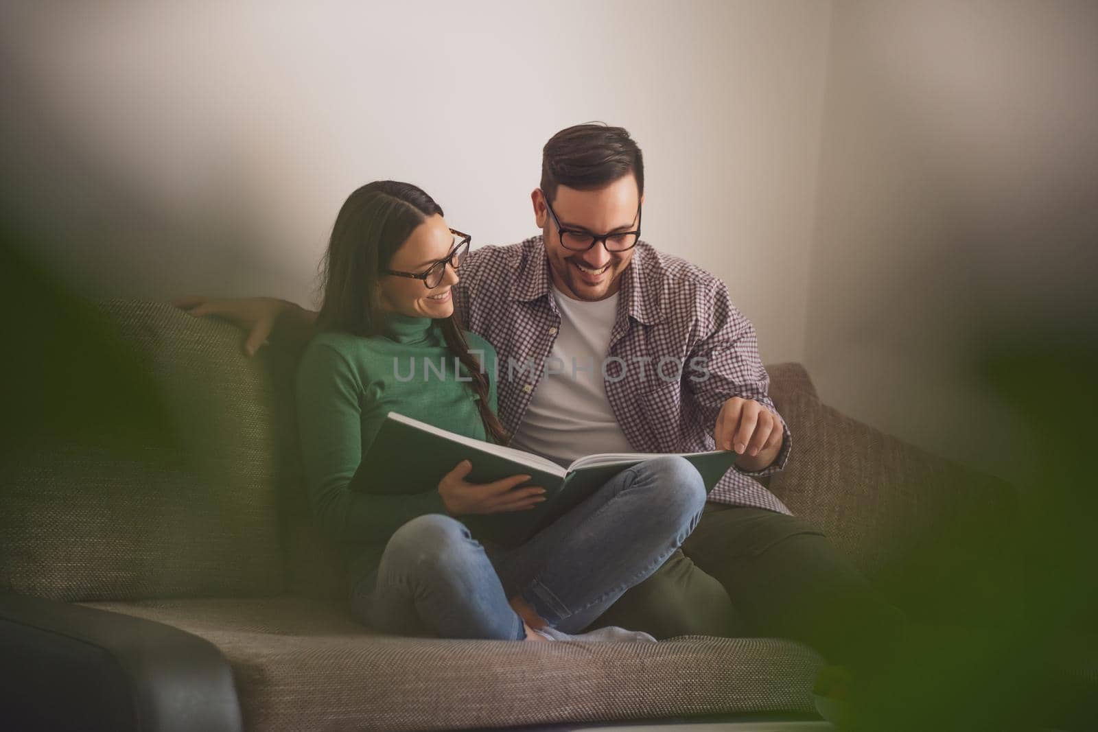 Happy couple is sitting at sofa in their home and looking at book.