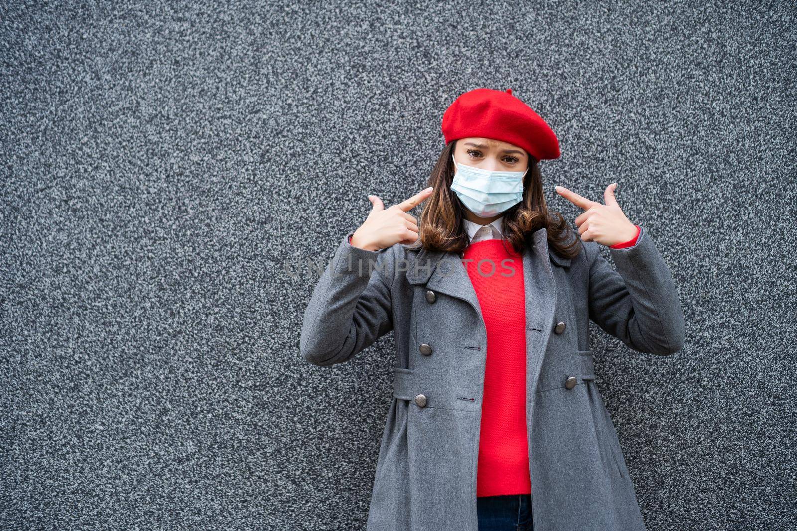 Adult woman in modern casual clothing standing in front of gray background. She is pointing at protective mask. Copy space for your advert.