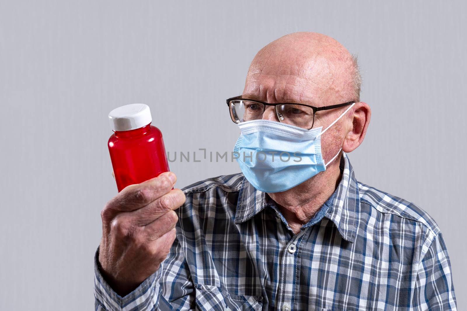 Old bald man with glasses and medical mask with red jar of pills in his hand. Copy space. by Essffes