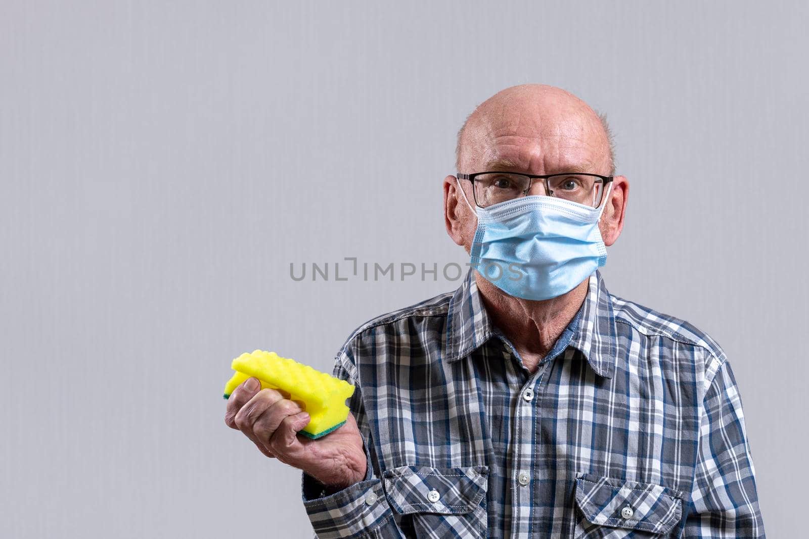 Old bald man with glasses and medical mask with sponge for washing dishes in his hand. Copy space. by Essffes