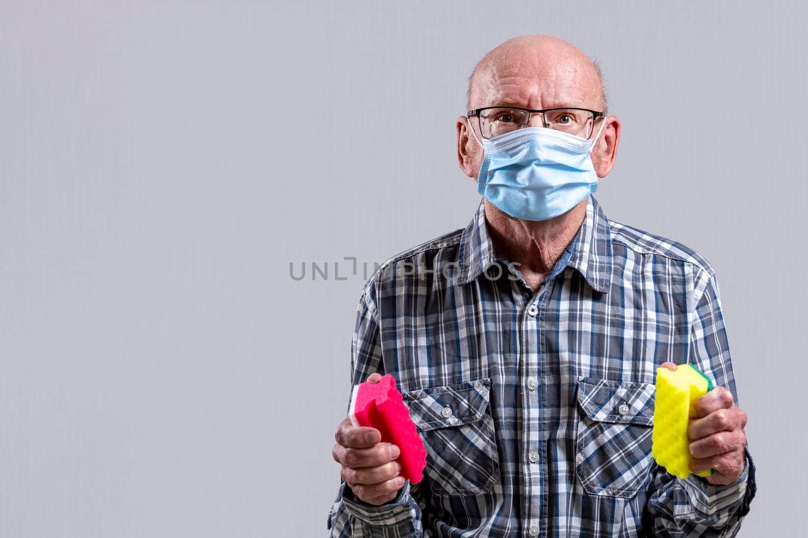 Old bald man with glasses and medical mask with sponges for washing dishes in his hands. Copy space. by Essffes