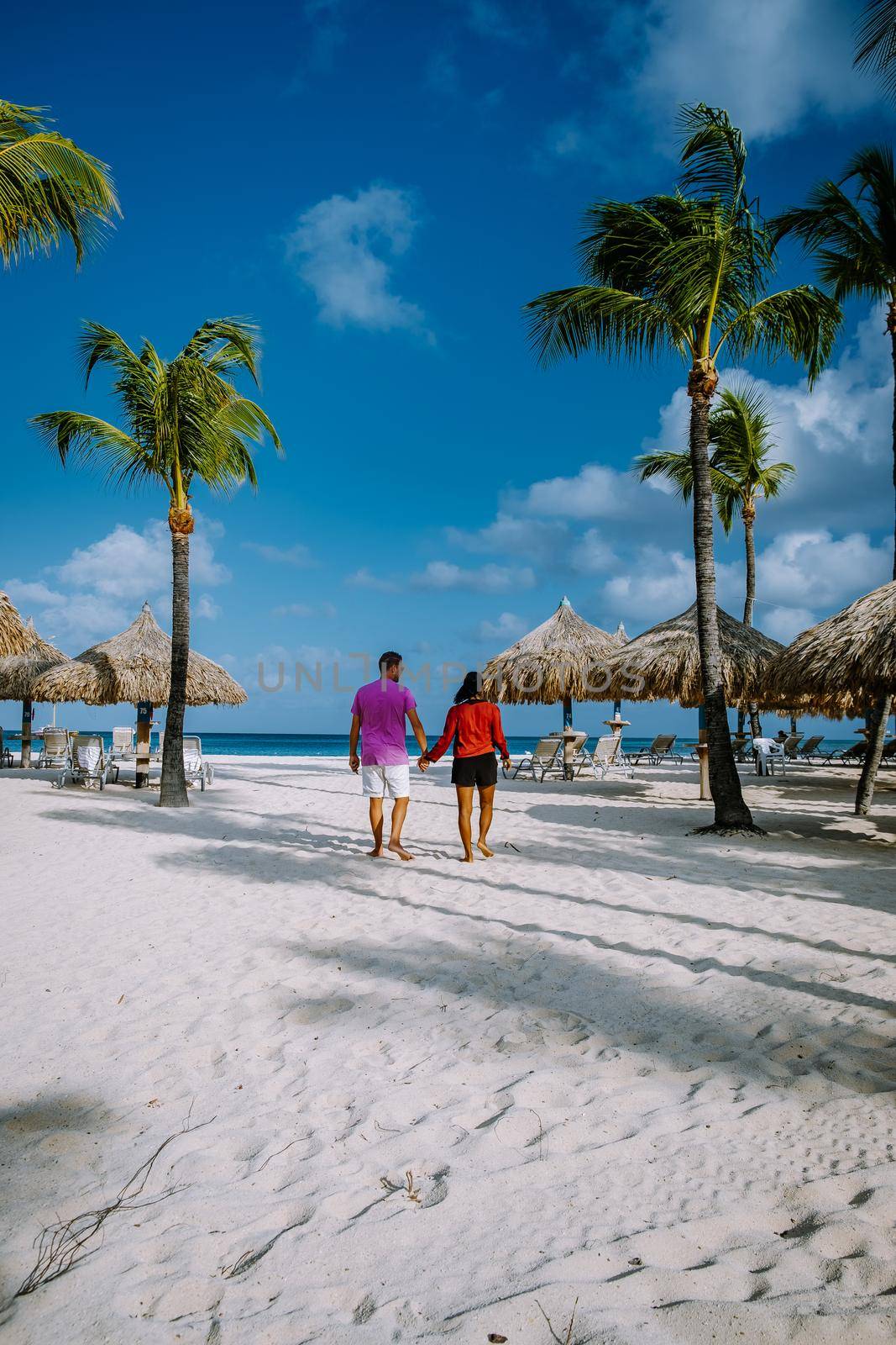Palm Beach Aruba, Amazing tropical beach with palm trees entering the ocean against azur ocean, gold sand, and blue sky. man and woman walking at a white beach, mid age couple