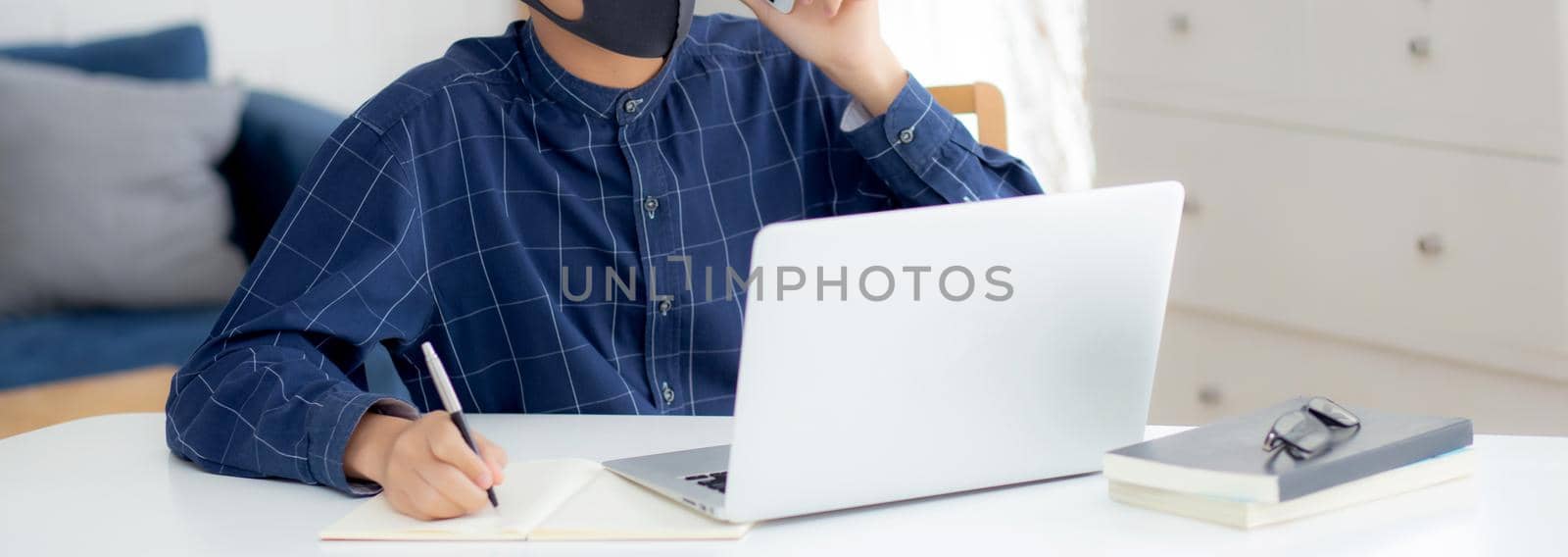 Young asian business man in face mask talking smartphone and work on laptop computer for protect covid-19, businessman quarantine work from home and writing notebook, social distancing and stay home.