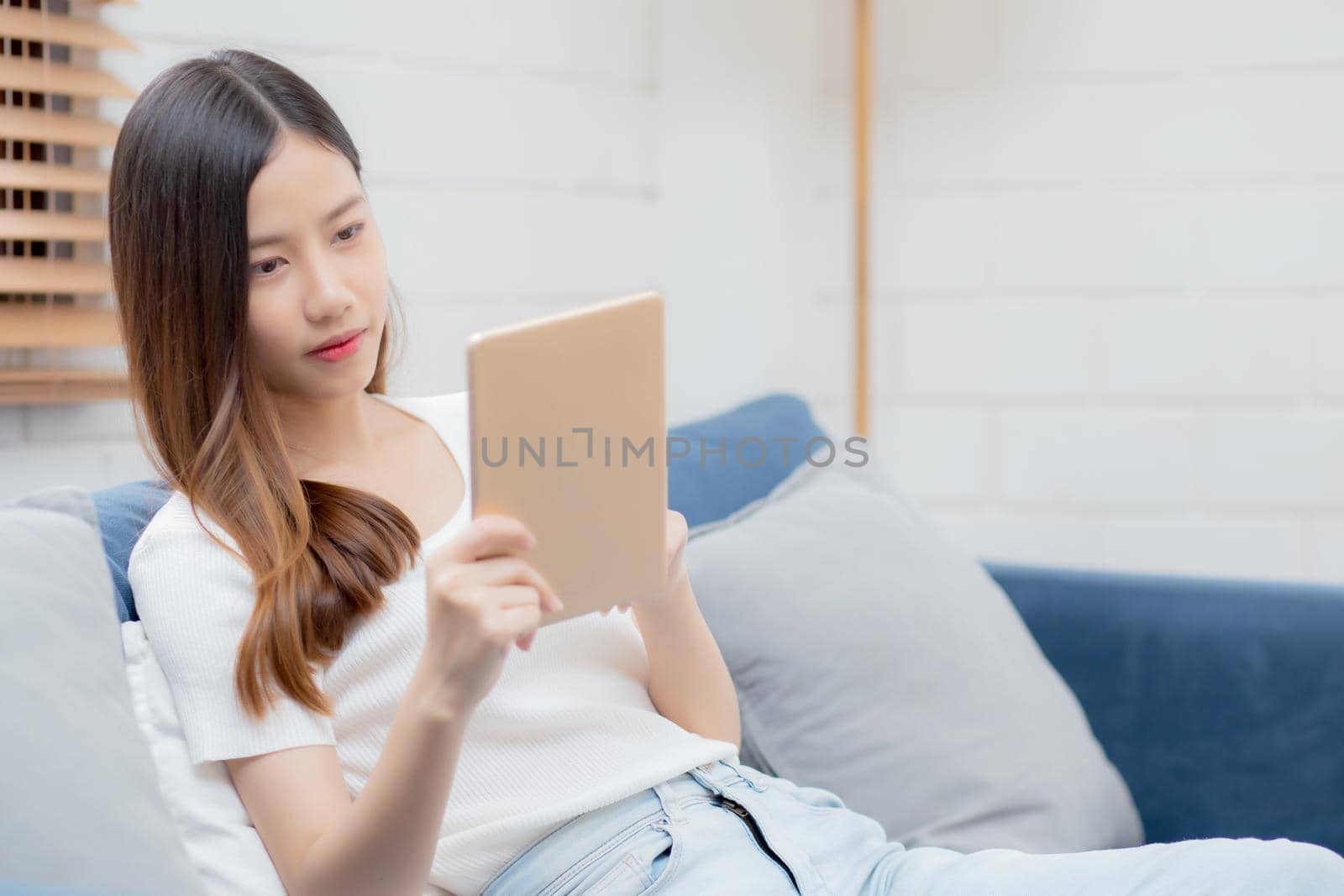 Young attractive asian woman resting using browsing tablet computer on sofa at home, happy girl sitting on couch relax reading digital gadget at house, communication and lifestyle concept.