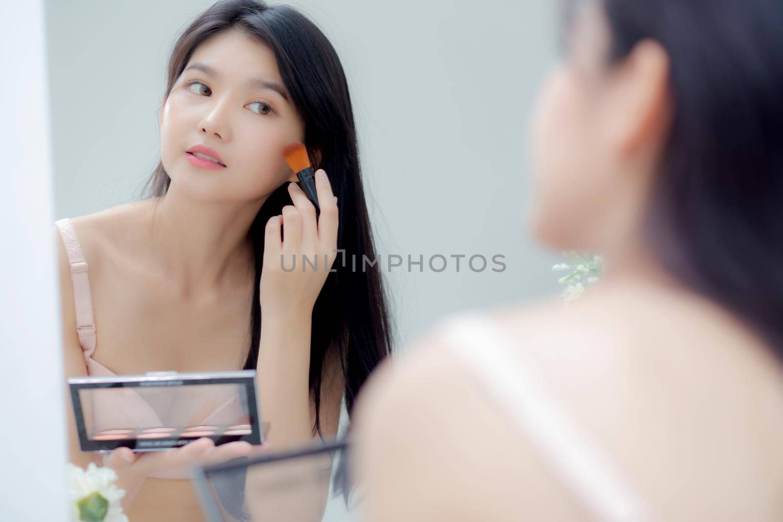Beauty young asian woman happy and smile with face looking mirror applying makeup with brusher of cheek in the room, beautiful of girl holding brush make-up on facial, skin care and cosmetic concept.