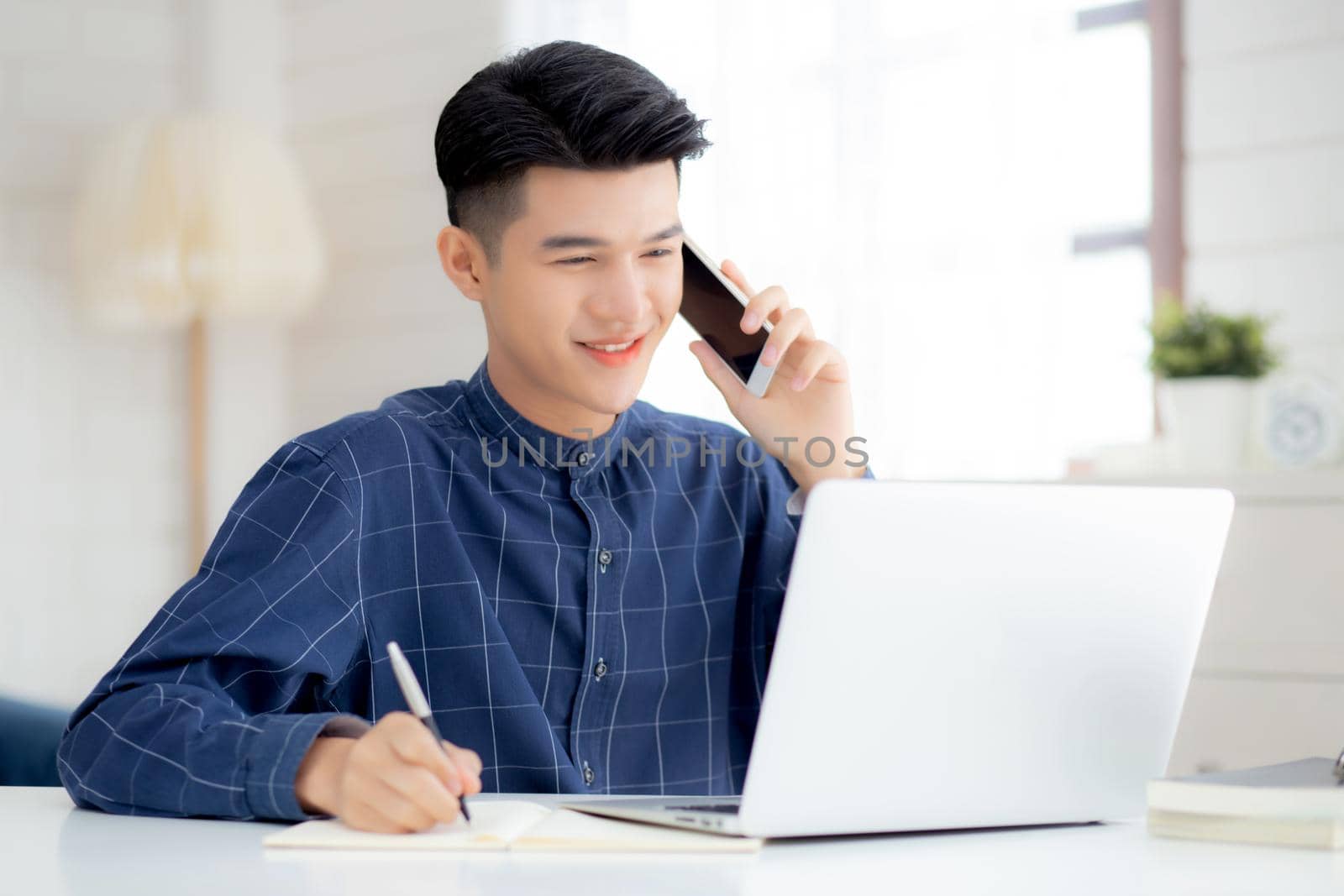 Young asian man talking phone and work from home with laptop computer, freelance male writing on notebook and speak on smartphone, stay home, domestic life, business and communication concept.