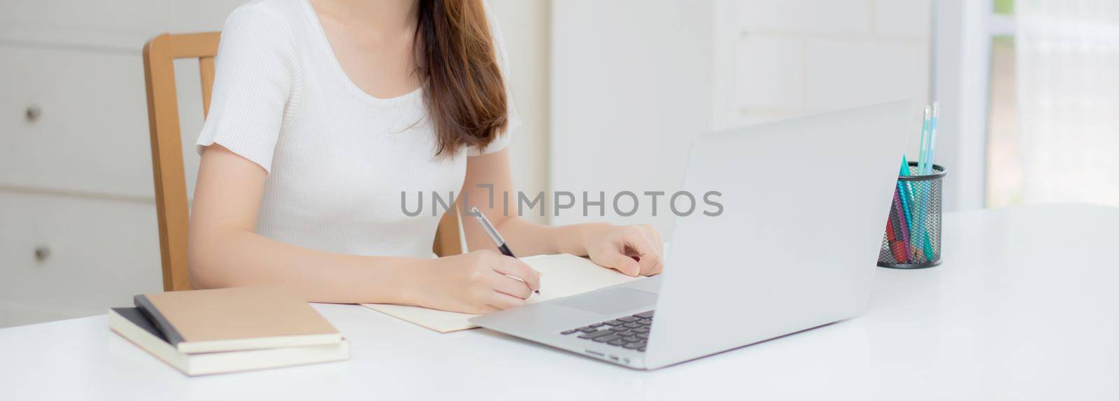 Young asian woman writing on notebook for planning working and using laptop computer on desk at home, girl notes about finance, female study and learning, business and communication concept.