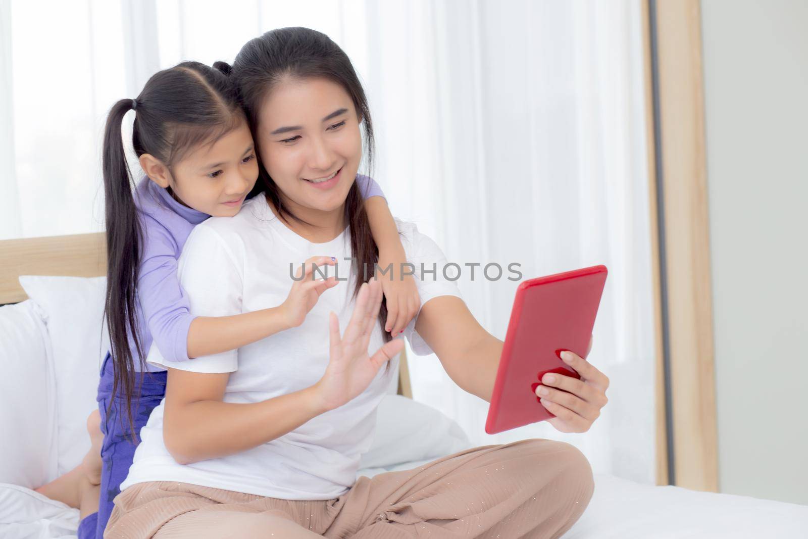 Young asian mom and daughter video call on tablet computer greeting with family together, happy mother and girl with relationship using technology in communication to internet online, two people. by nnudoo