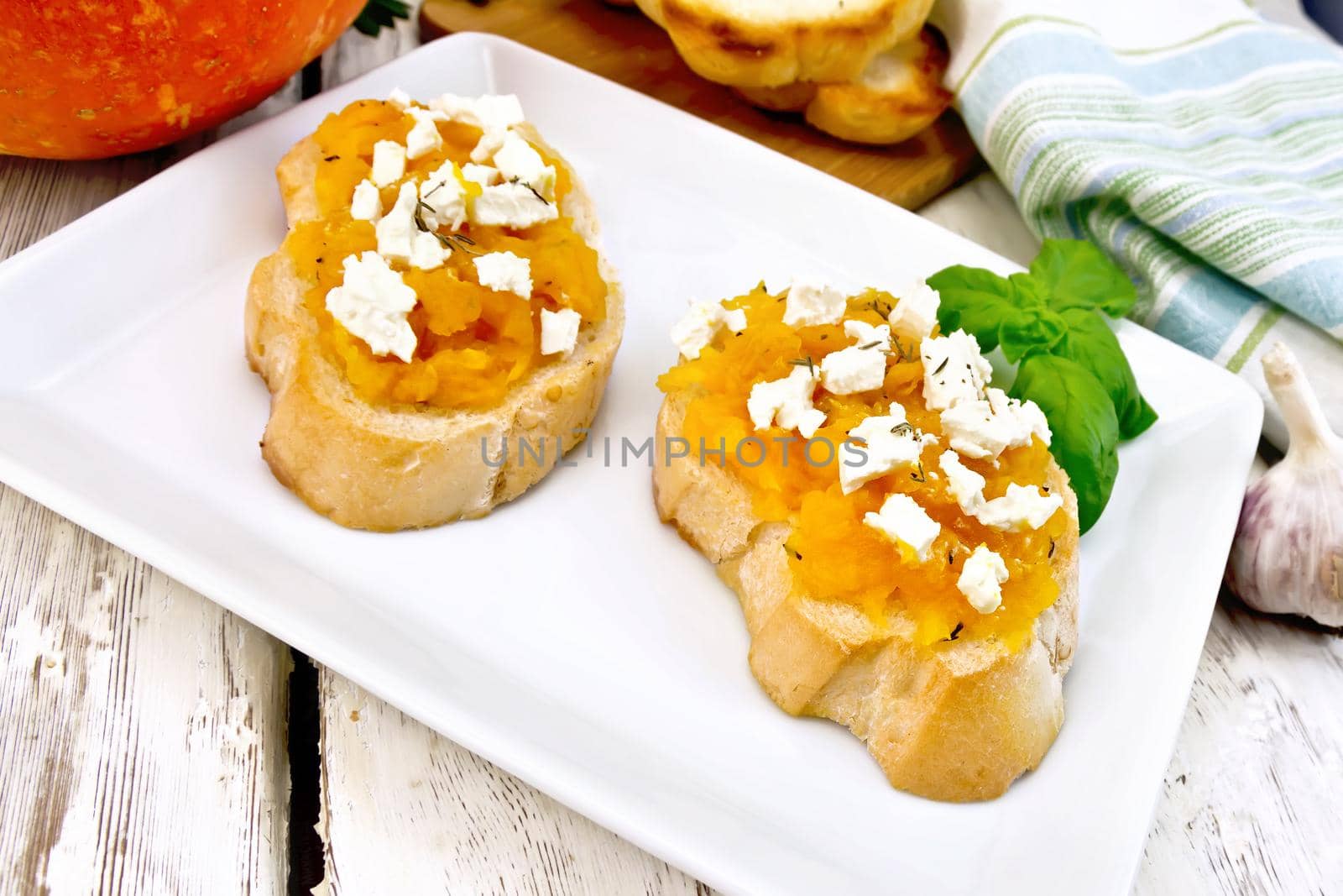 Bruschetta with pumpkin, salted feta cheese in a plate, garlic, basil and parsley, towel and vegetable on a light wooden board background