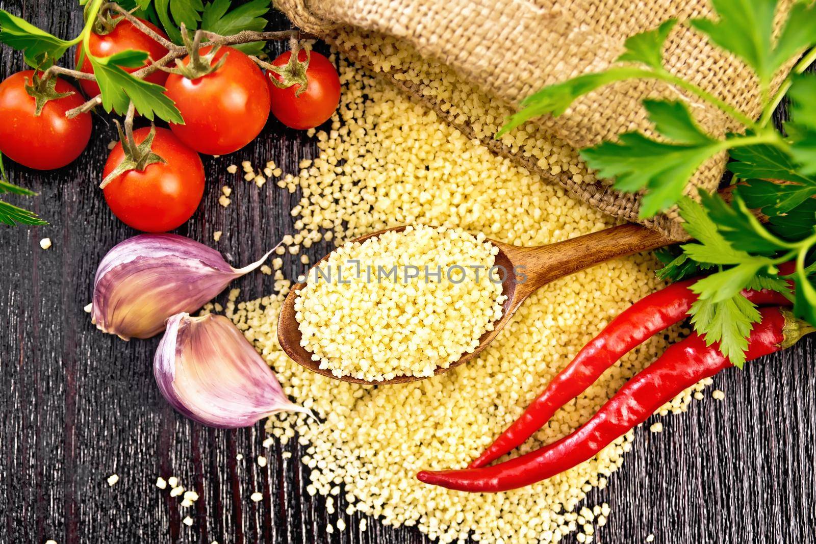 Raw couscous in a spoon and a sack of burlap, tomatoes, hot peppers, herbs and garlic on wooden board background from above