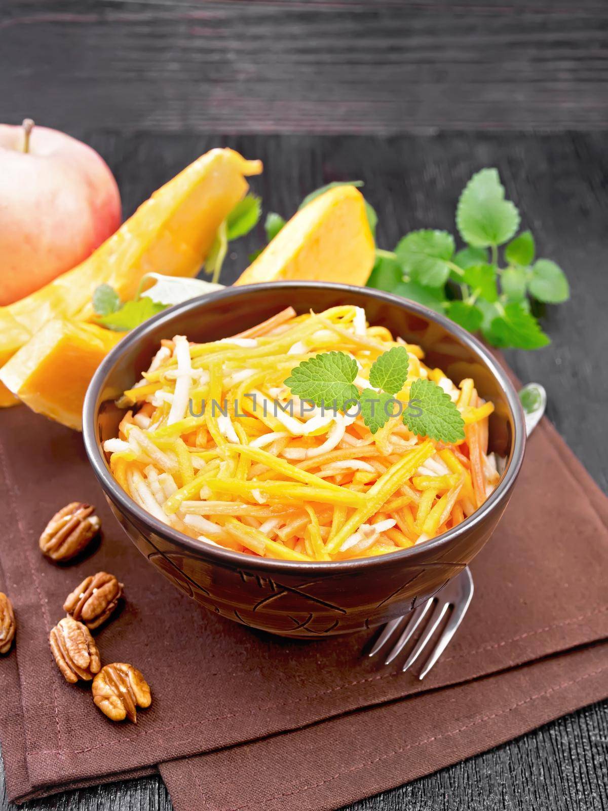 Pumpkin, carrot and apple salad with pecans seasoned with vegetable oil in a bowl on a napkin, mint on black wooden board background