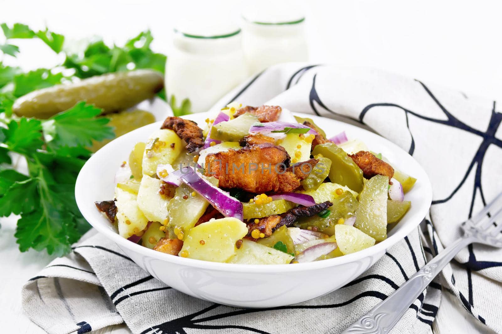 Salad of boiled potatoes, fried bacon, red onions and pickled cucumbers, seasoned with grain mustard, spices and vegetable oil in a plate on a napkin against white wooden board background