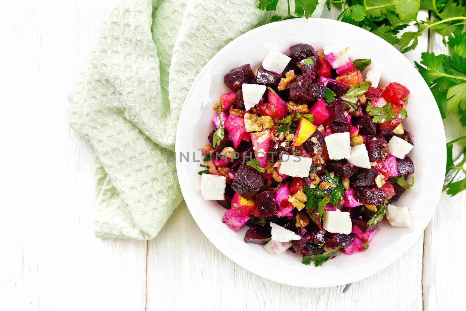 Salad with beetroot, feta cheese, apple, walnuts, parsley, seasoned with balsamic vinegar and olive oil in a plate, napkin against the background of a light wooden board on top
