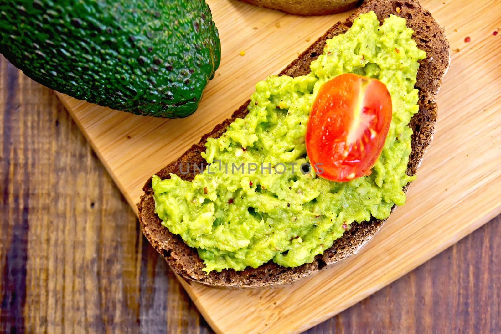 A slice of rye bread with guakomole and tomatoes, avocado on a wooden boards background