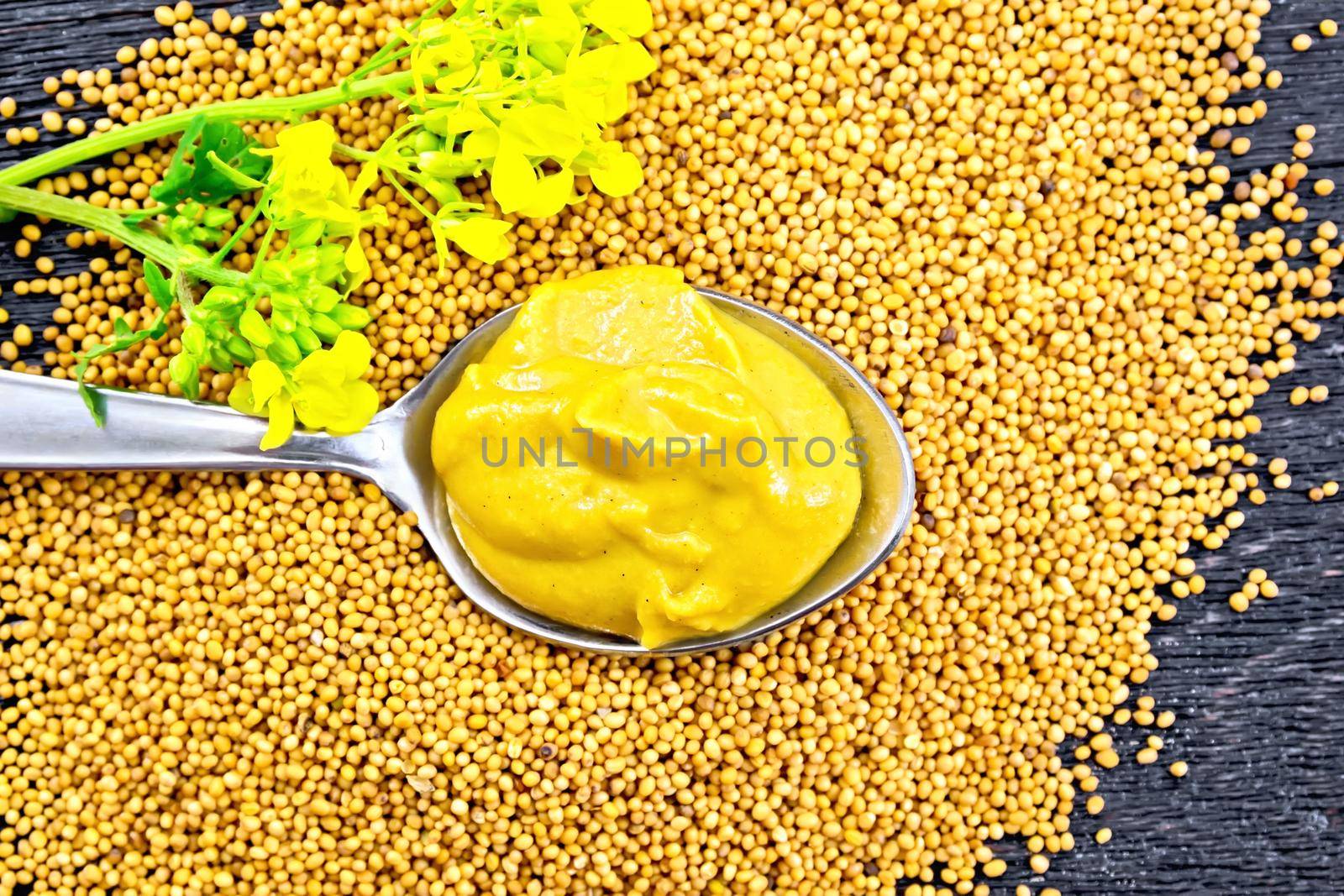 Mustard sauce in a metal spoon with a yellow flower on mustard seeds against the background of a wooden board from above