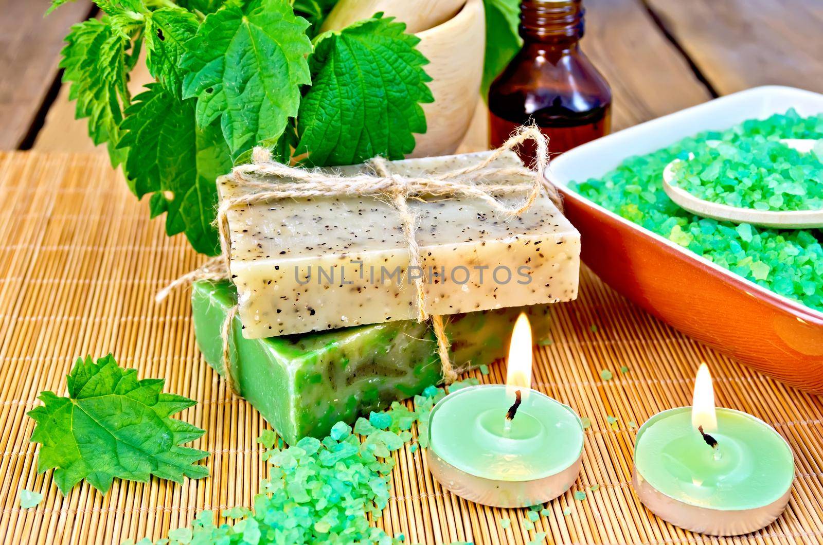 Two bars of homemade soap with twine, bath salt in a bowl, oil in the bottle, nettles in a mortar, two candles on a wooden boards background