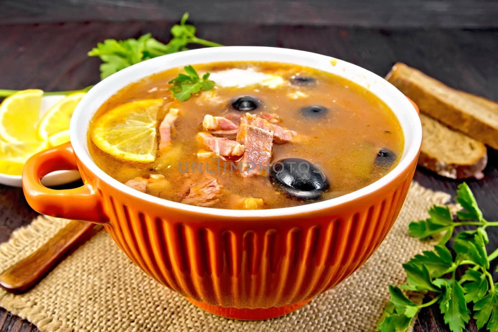 Soup saltwort with lemon, meat, pickles, tomato sauce olives in a bowl on a sacking, bread and parsley on a wooden board background