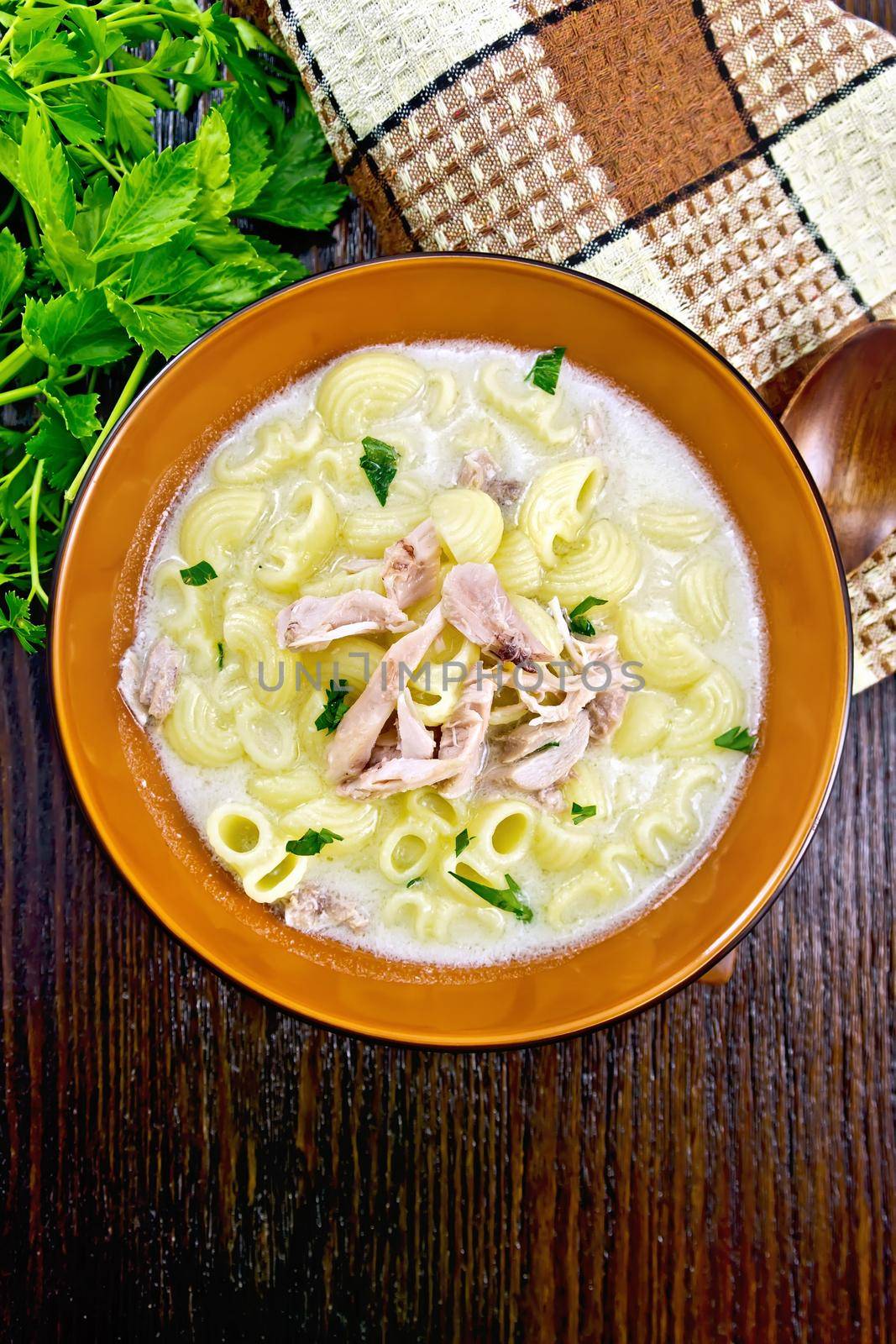 Chicken meat soup, pasta with cream and cilantro in a clay plate, towel, parsley, spoon on a wooden board background from above