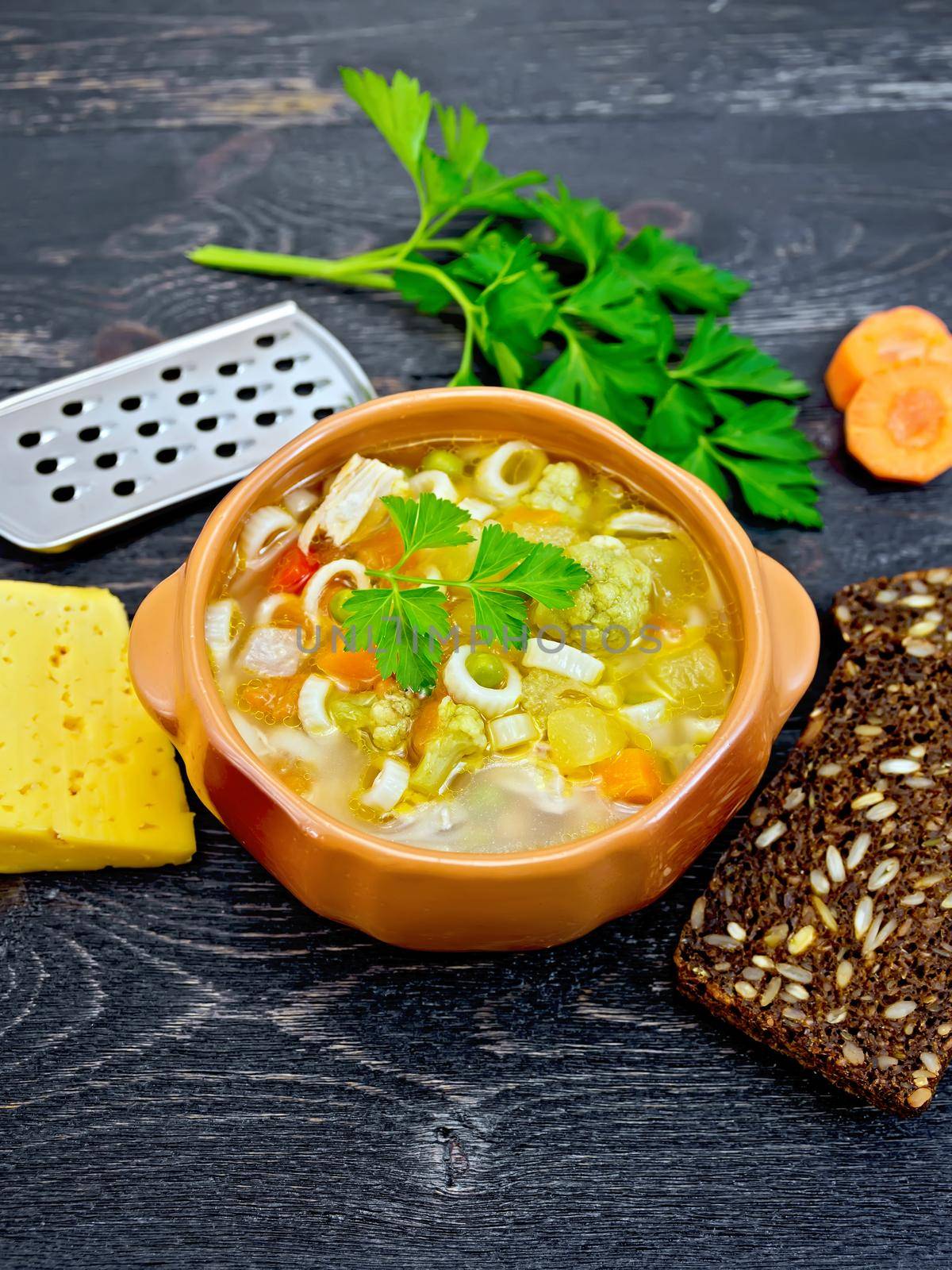 Minestrone soup with meat, celery, tomatoes, zucchini and cabbage, green peas, carrots and pasta in an earthenware bowl, cheese, grater on black background wooden plank
