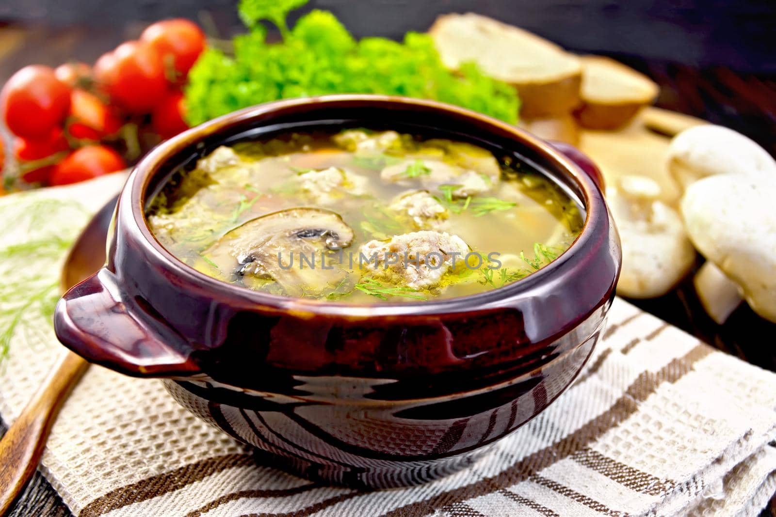 Soup with meatballs, noodles and champignon in a clay bowl, spoon on the towel, parsley, tomatoes, mushrooms and bread on a wooden boards background