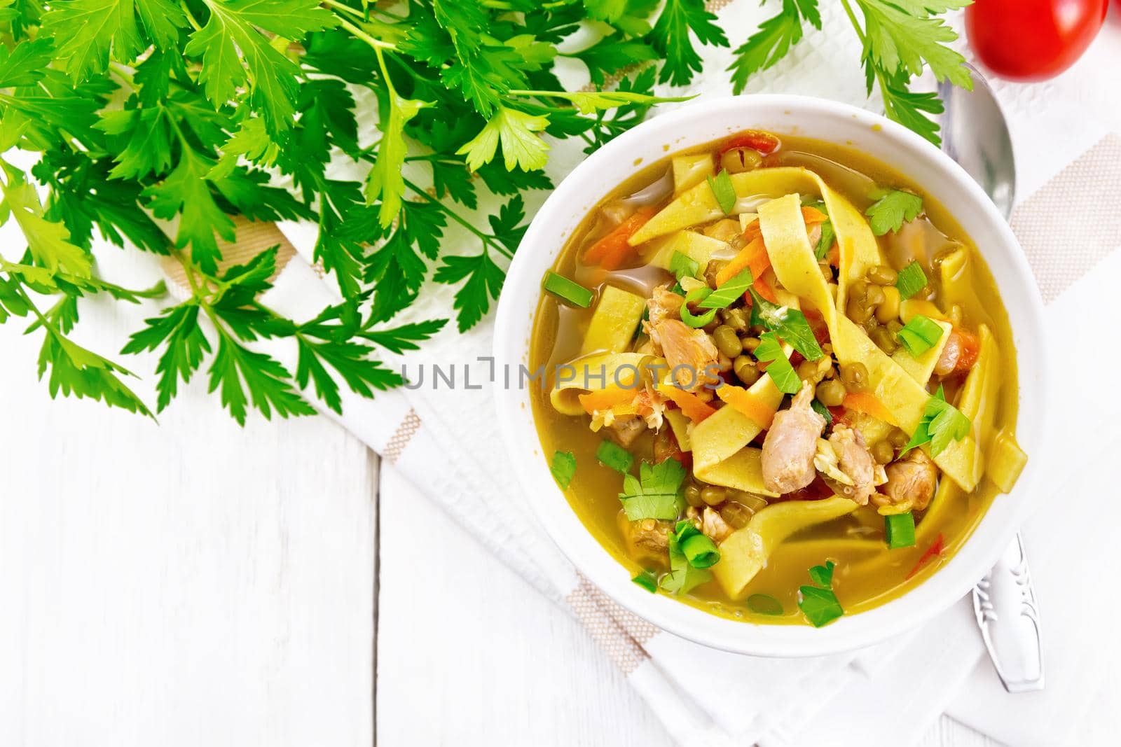 Soup with meat, tomatoes, vegetables, mung bean lentils and noodles in a bowl on napkin, parsley and a spoon on the background of light wooden board from above