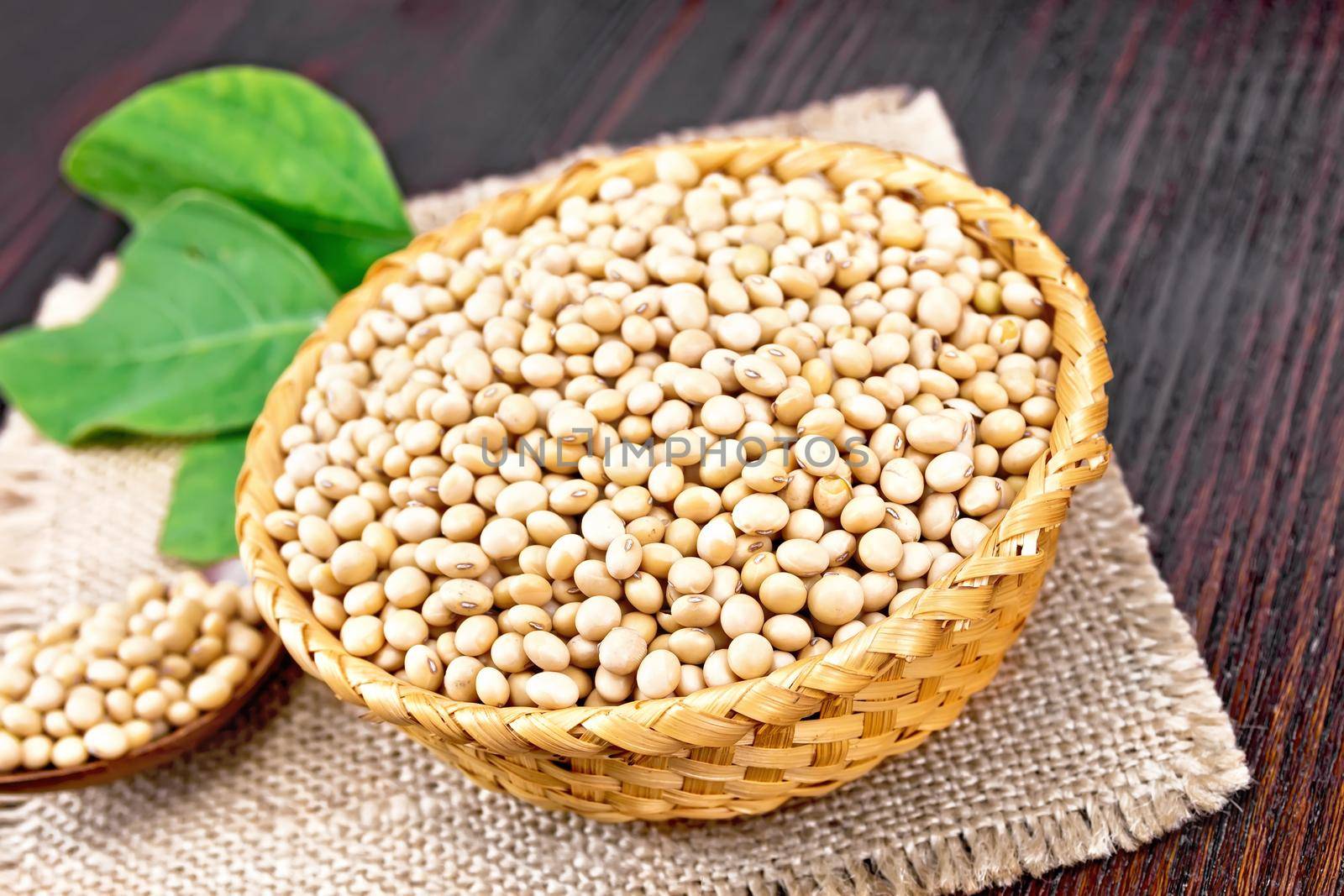 Soy beans in wicker bowl and spoon, green leaf on burlap napkin on wooden board background