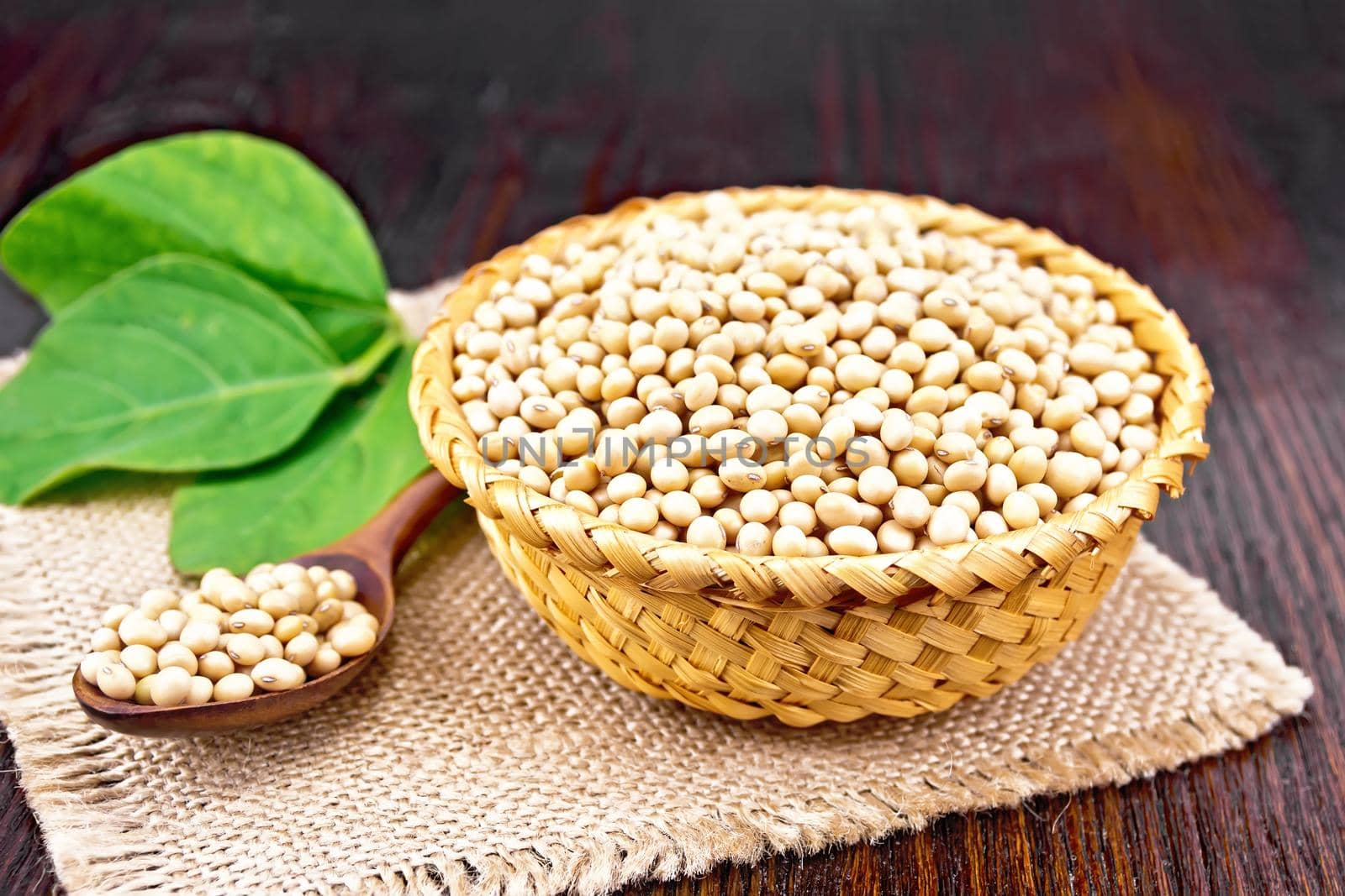 Soy beans in wicker bowl and spoon, green leaf on burlap on wooden board background