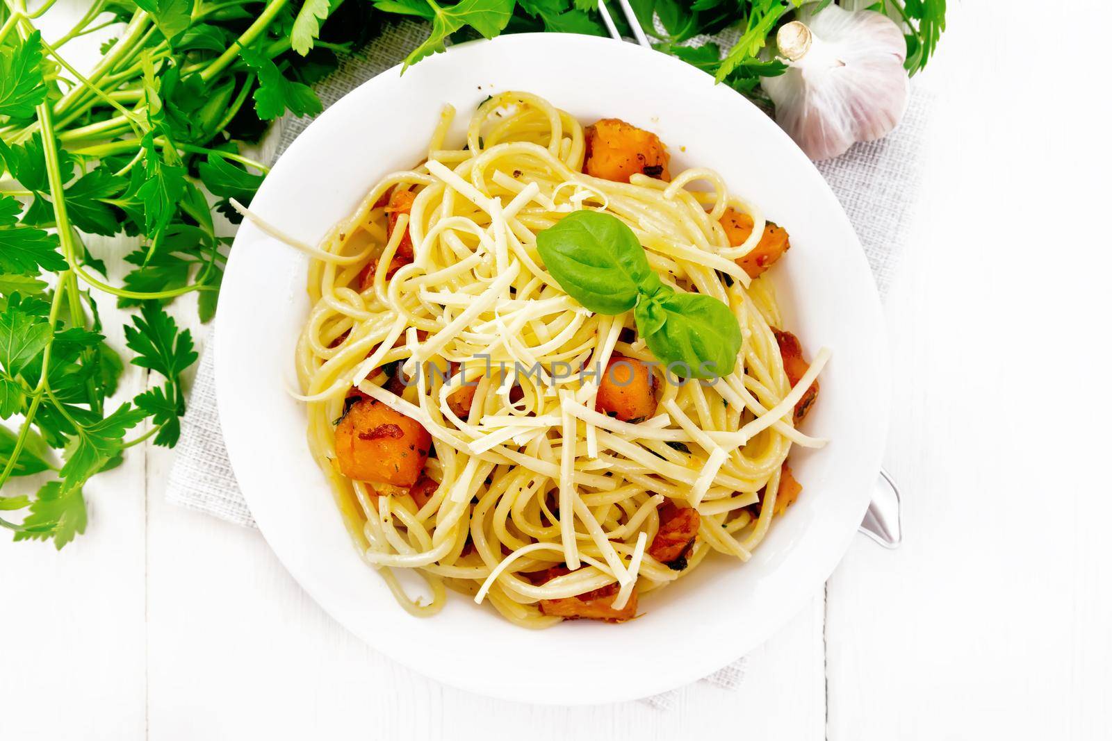 Spaghetti with pumpkin, onion and garlic in a plate on a napkin, basil, parsley and fork on wooden board background from above