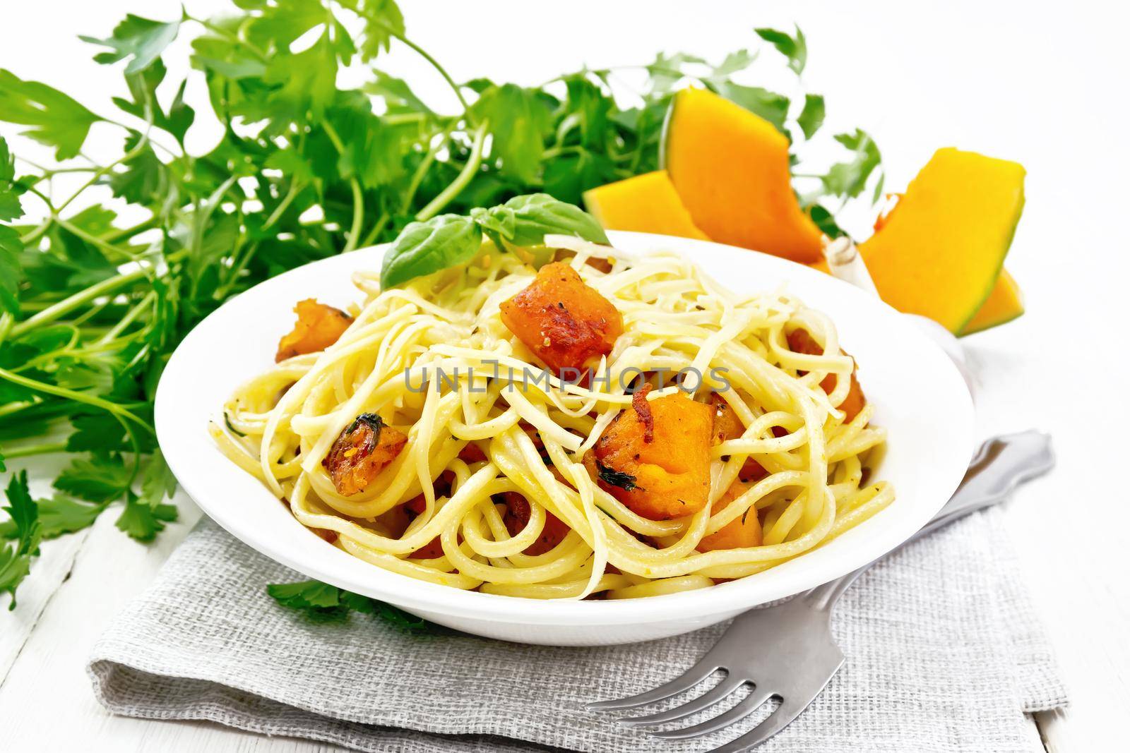 Spaghetti with pumpkin, onion and garlic in plate on a towel, basil, parsley and a fork on wooden board background