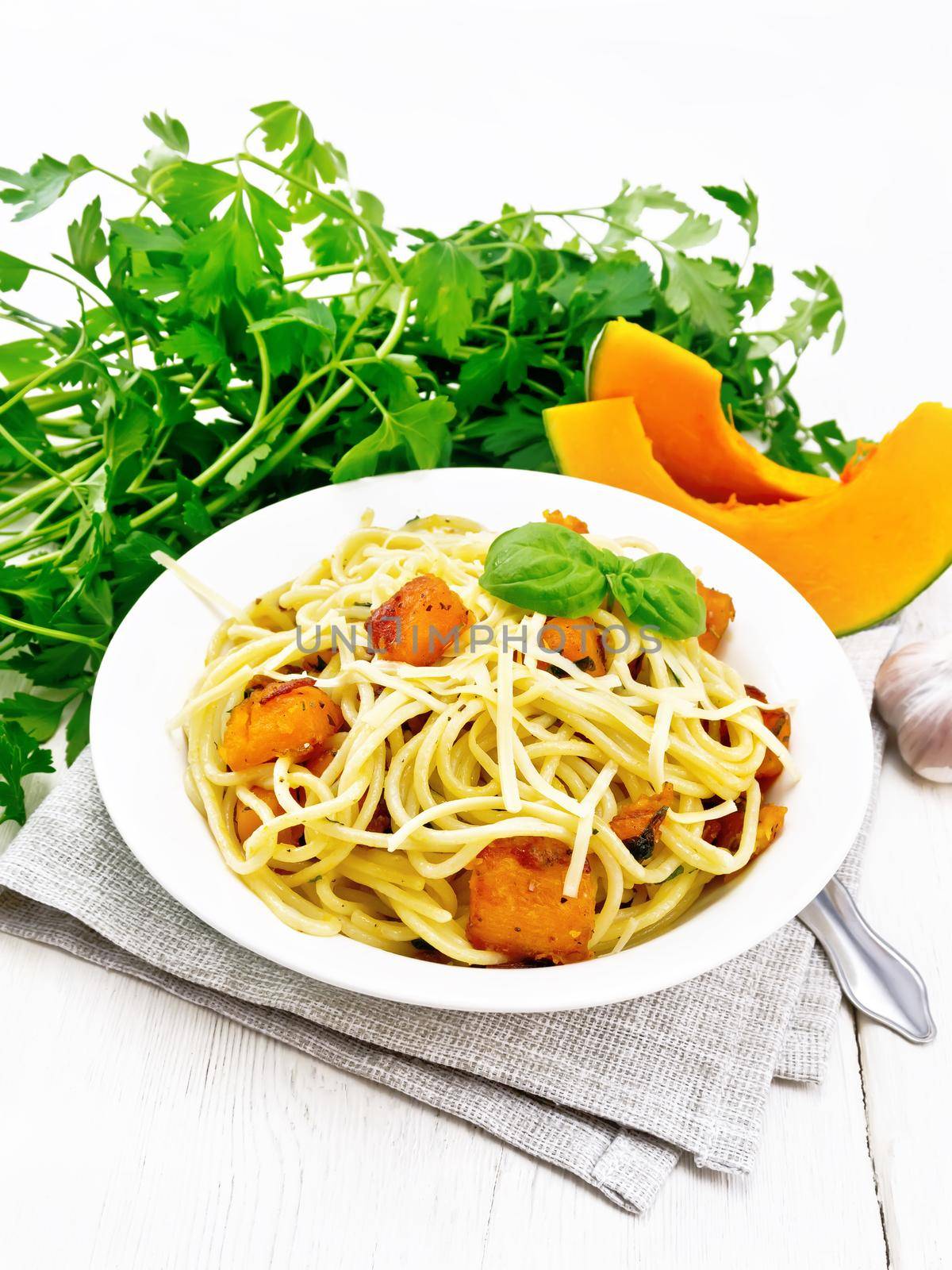 Spaghetti with pumpkin, onion and garlic in a plate on a napkin, basil, parsley and fork on wooden board background