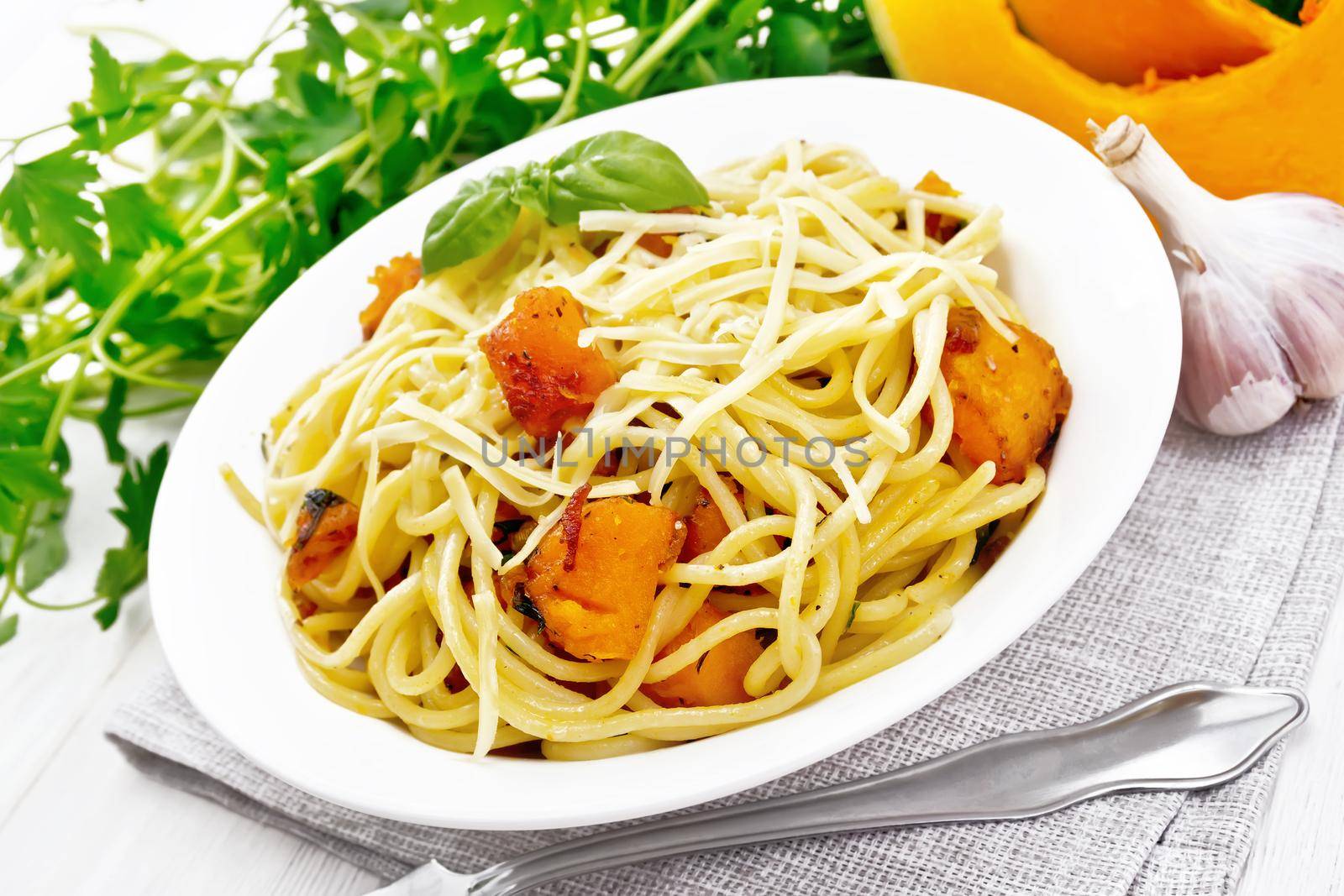 Spaghetti with pumpkin, onion and garlic in a plate on a napkin, basil, parsley and fork on white wooden board background
