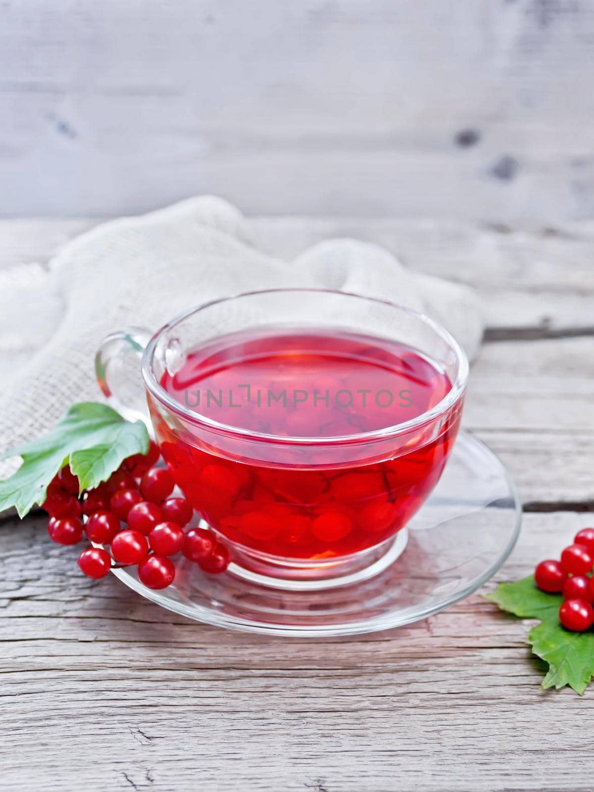 Tea with viburnum in a glass cup, berries and green leaves, burlap napkin on wooden board background