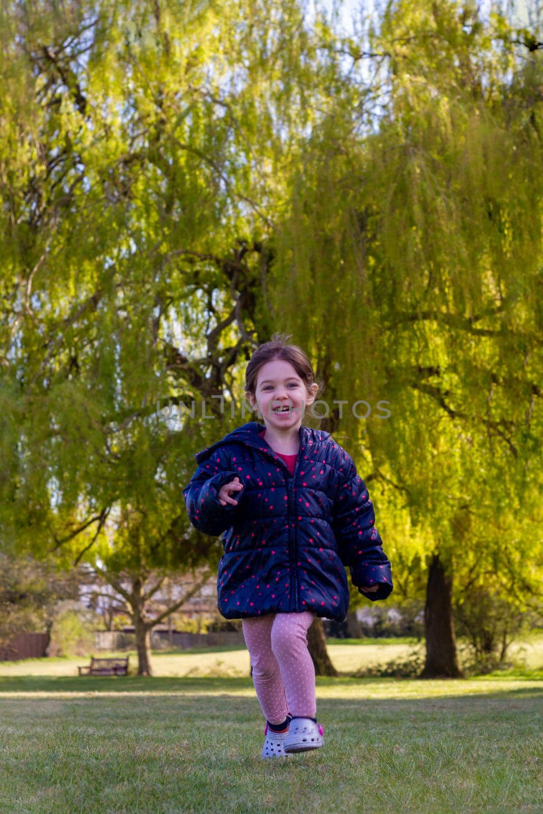 Full-length shot of a cute brown hair baby girl with a blue jacket and pink trousers running on the grass in a park on a sunny spring day