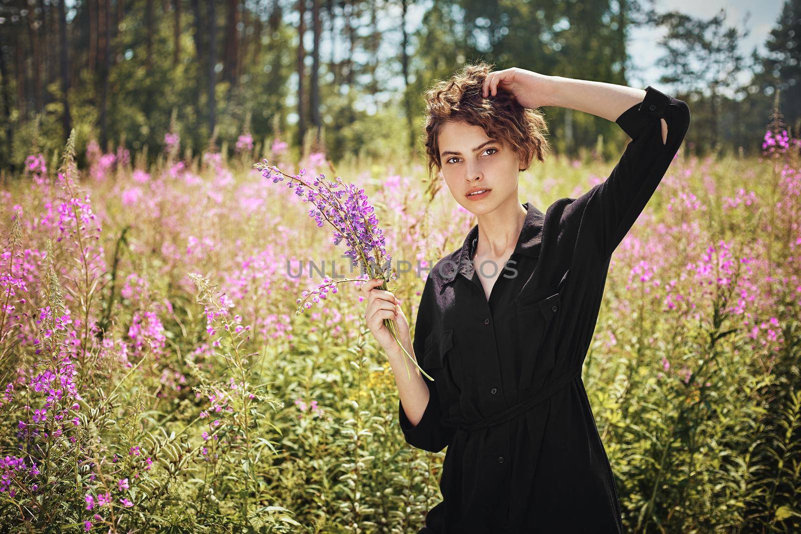 Young woman with a bouquet of lupines in her hand against the background of blooming Ivan-tea on a bright sunny summer day