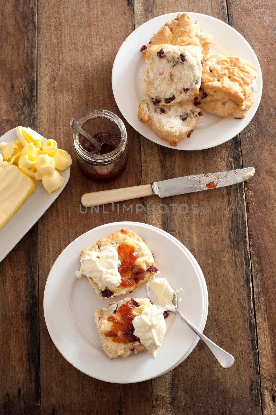 Freshly baked fruity rock cakes or raisin buns served with jam and whipped cream in an overhead view on a wooden table with pat of butter alongside