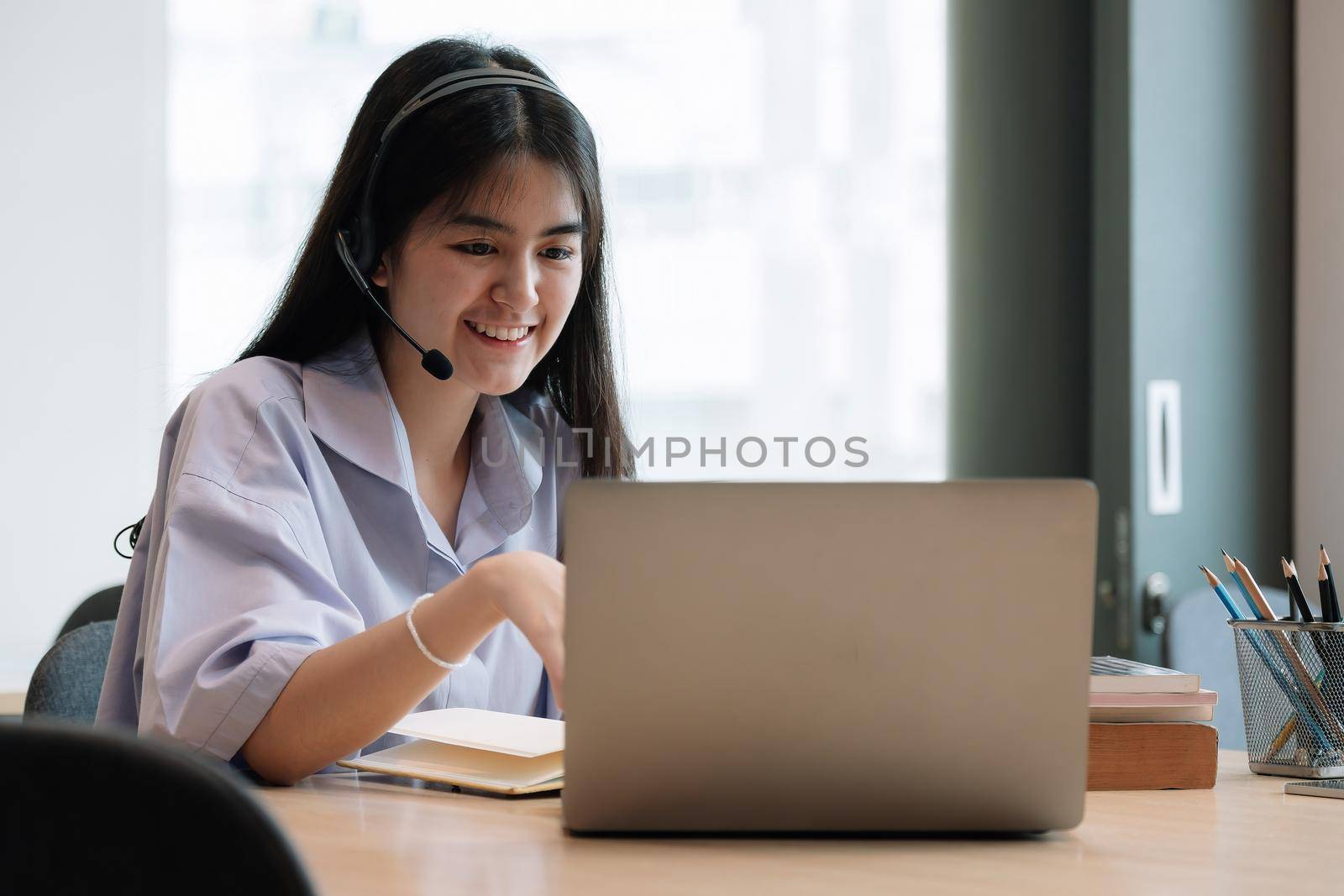 Asian young female student sitting while using headphones and laptop computer when studying online.