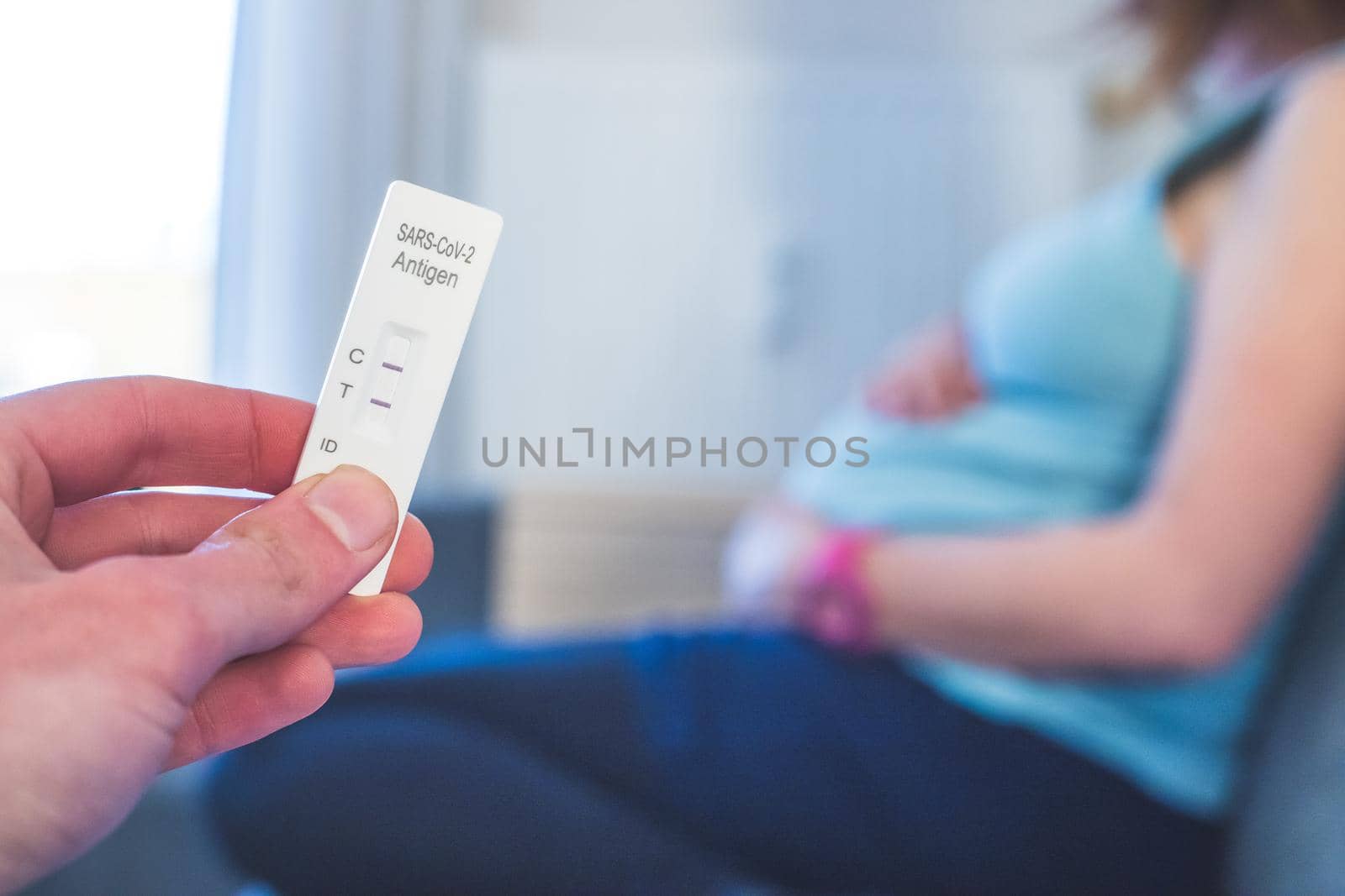 Close up of fingers holding express antigen covid test, positive result. Pregnant woman in the blurry background