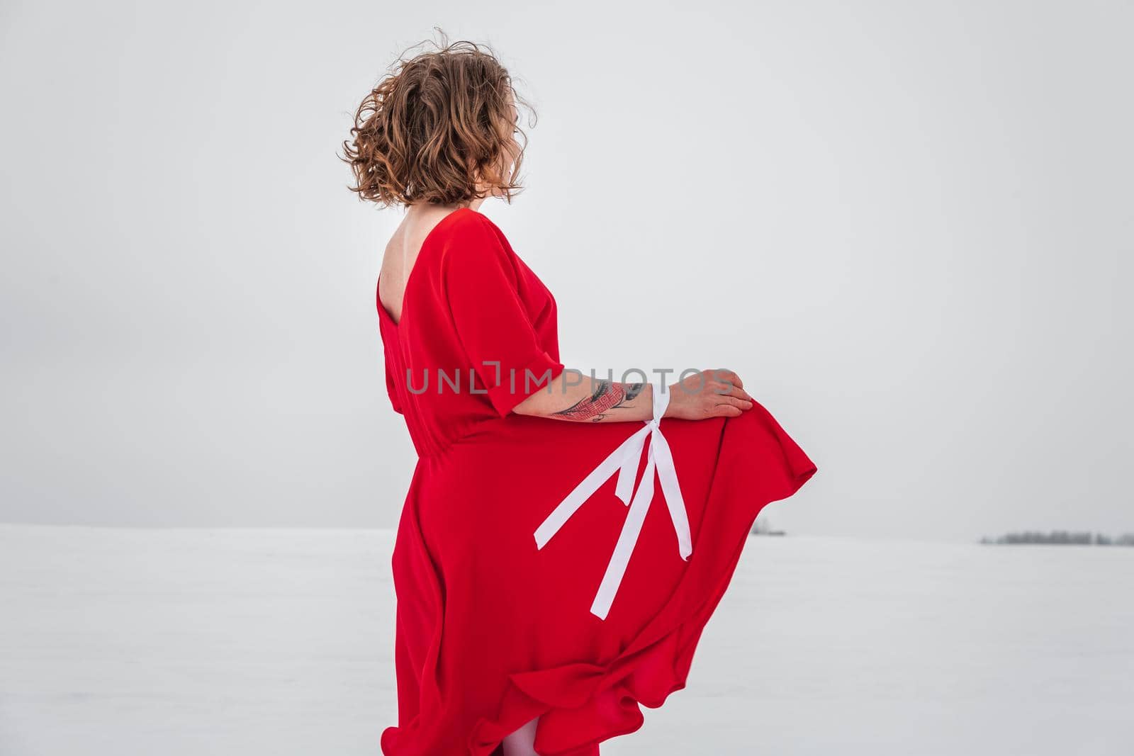 Beautiful woman in a red dress posing outside on a snowy field. She is wearing red dress and have tattoo on her arm.