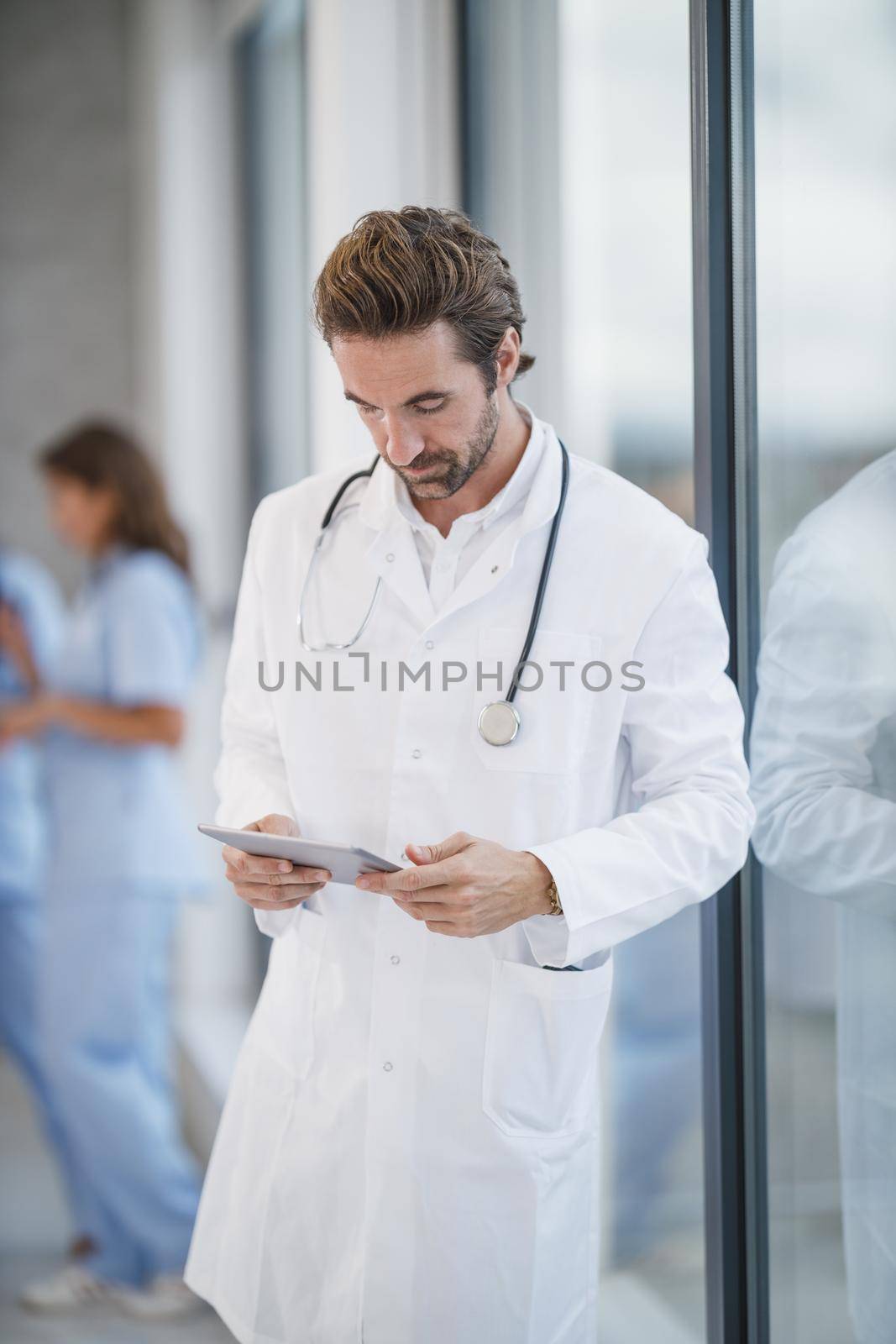 Shot of a mature doctor using digital tablet while standing near a window in a hospital hallway during the Covid-19 pandemic.