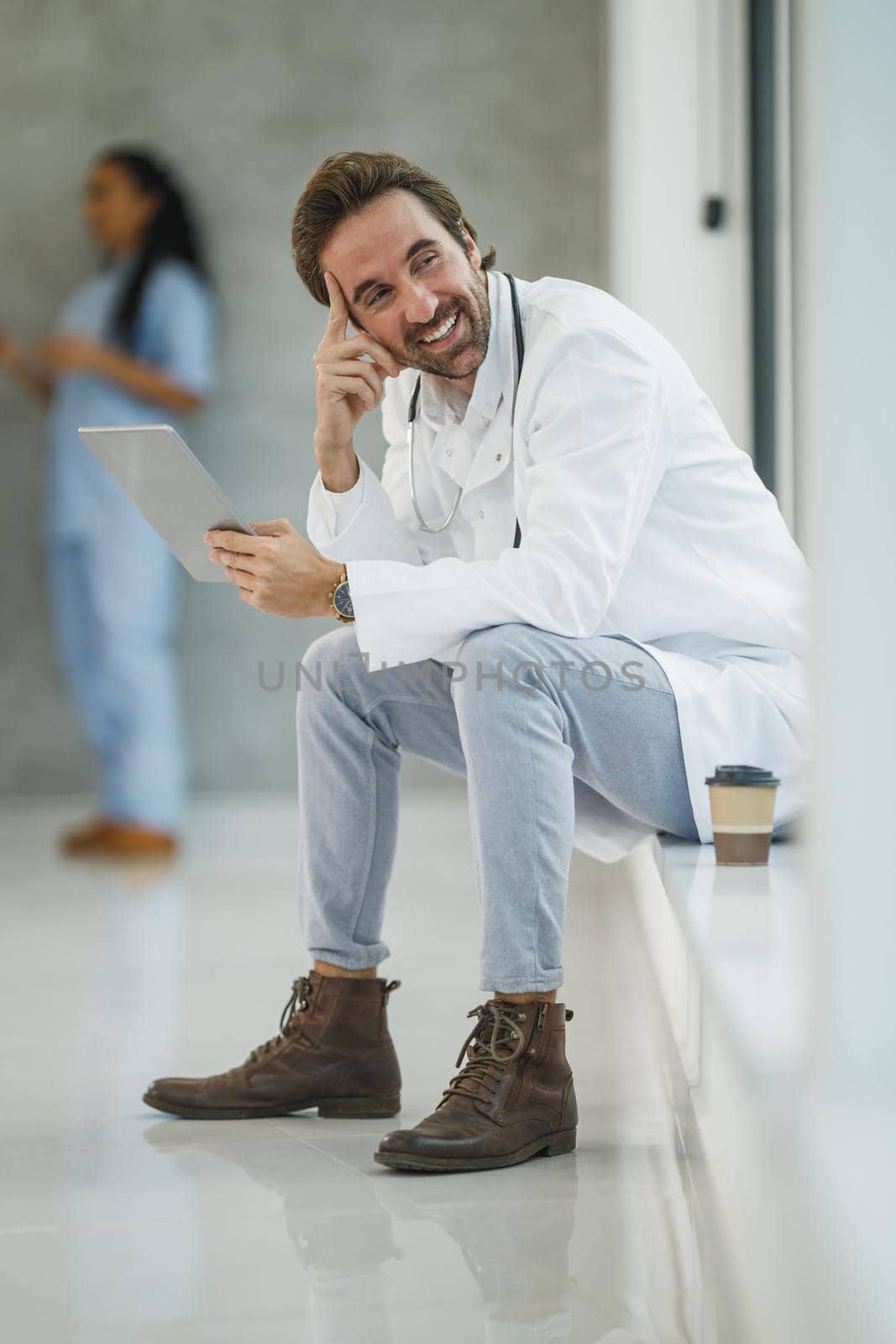 Shot of a smiling doctor using digital tablet while sitting near a window in a hospital hallway during the Covid-19 pandemic.