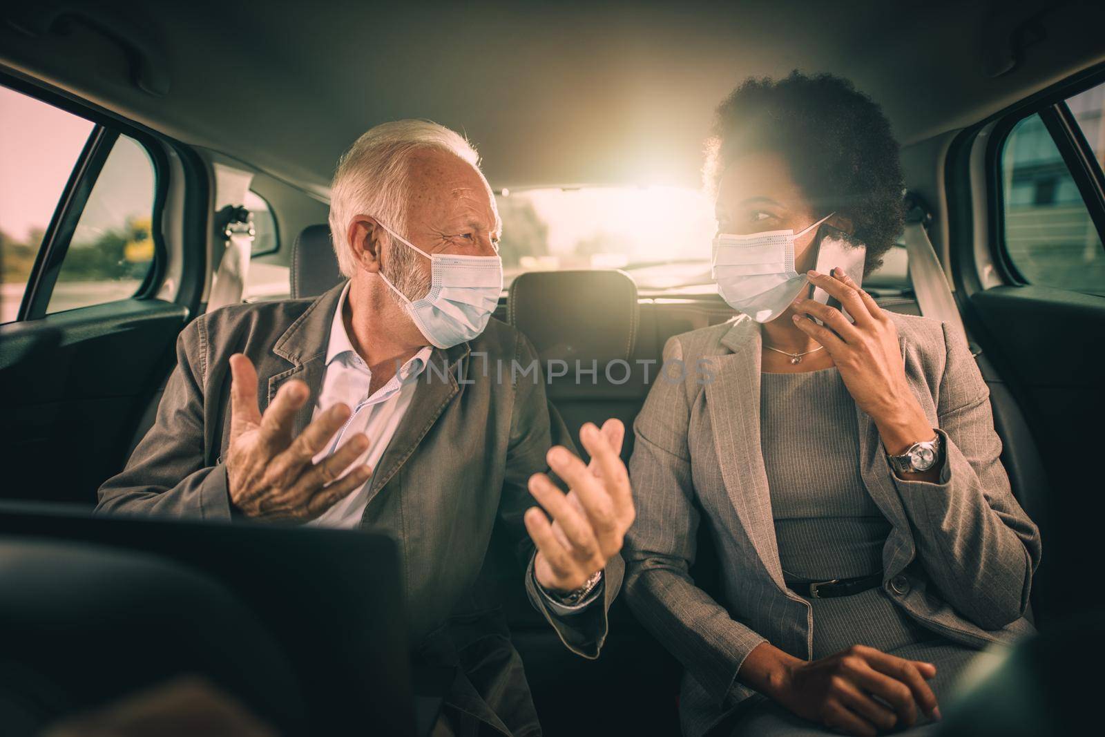 Shot of two successful multi-ethnic business people with protective mask talk about work while sitting in the backseat of a car on their morning commute during COVID-19 pandemic.