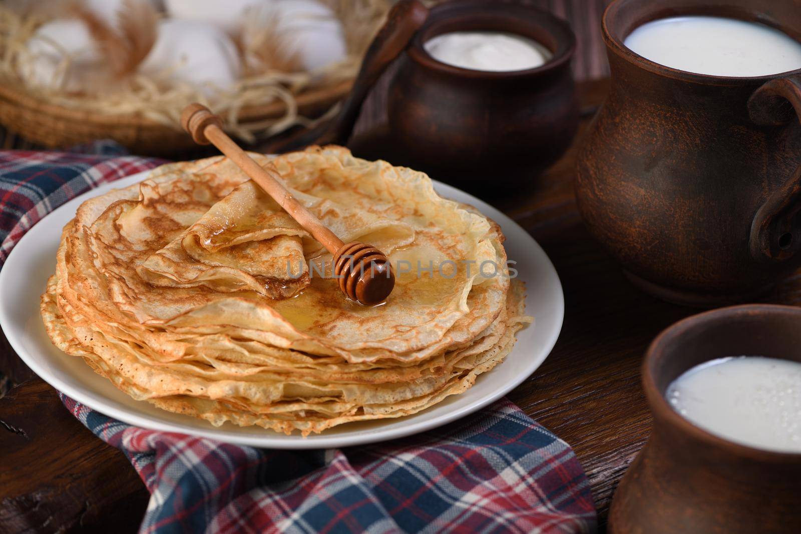 Homemade thin pancakes with honey stacked in a stack, on a wooden table with a mug of milk, a pot of sour cream and eggs in a basket. Traditional Slavonian, pagan holiday (Maslinitsa). Country style food.